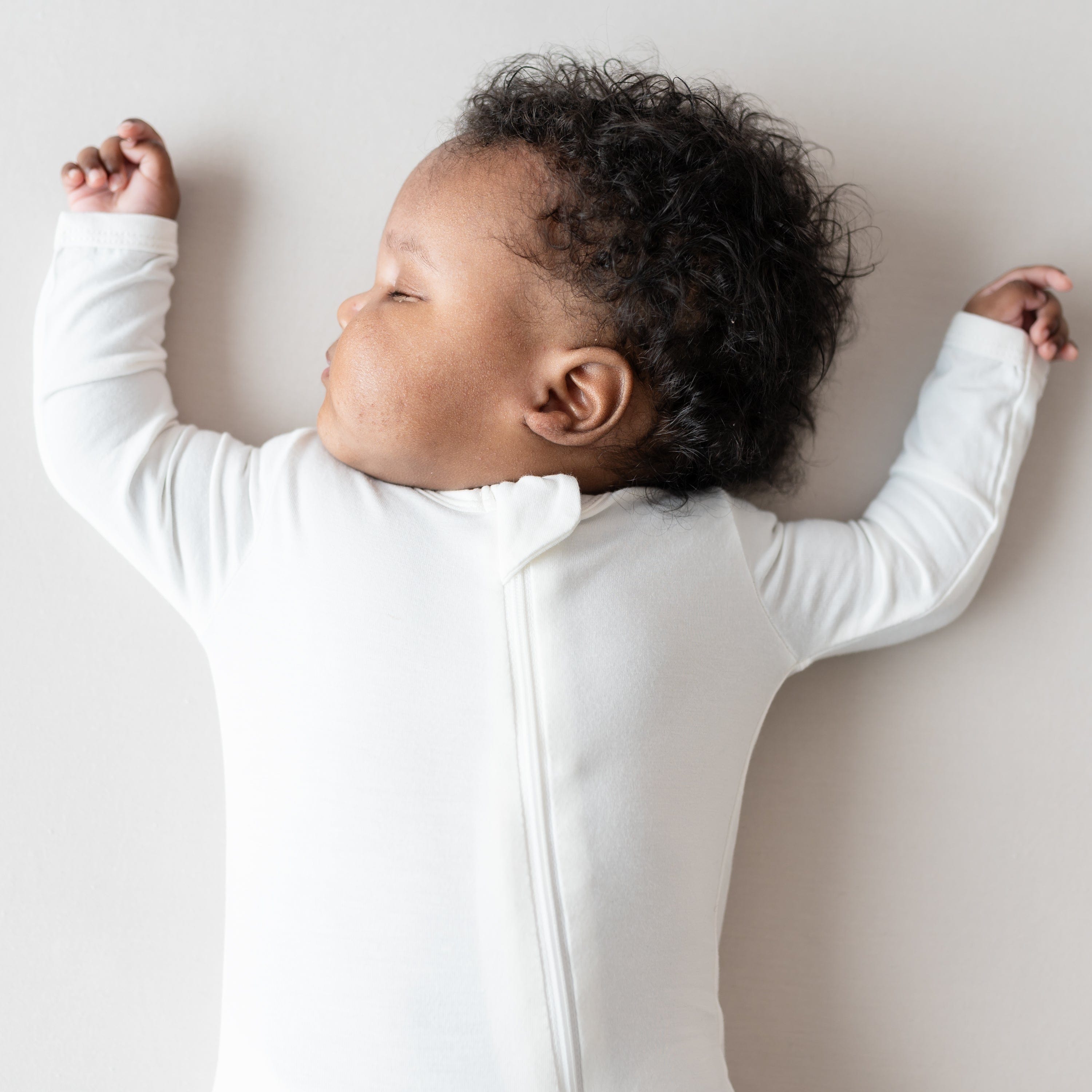 Close up of sleeping baby with both arms up while wearing a Kyte Baby Zippered Romper in Cloud