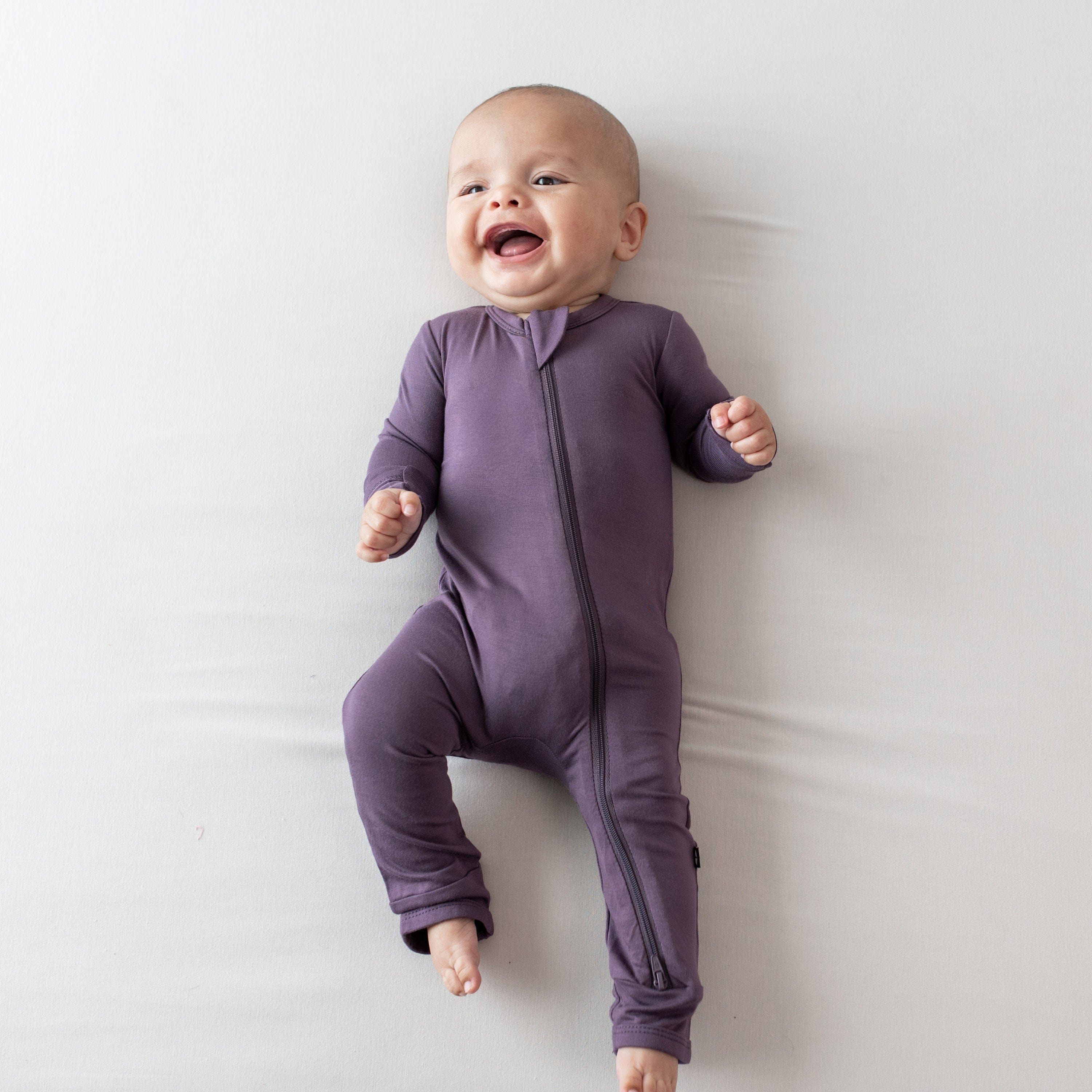 Smiling infant laying on an off-white surface wearing the Zippered Romper in Currant