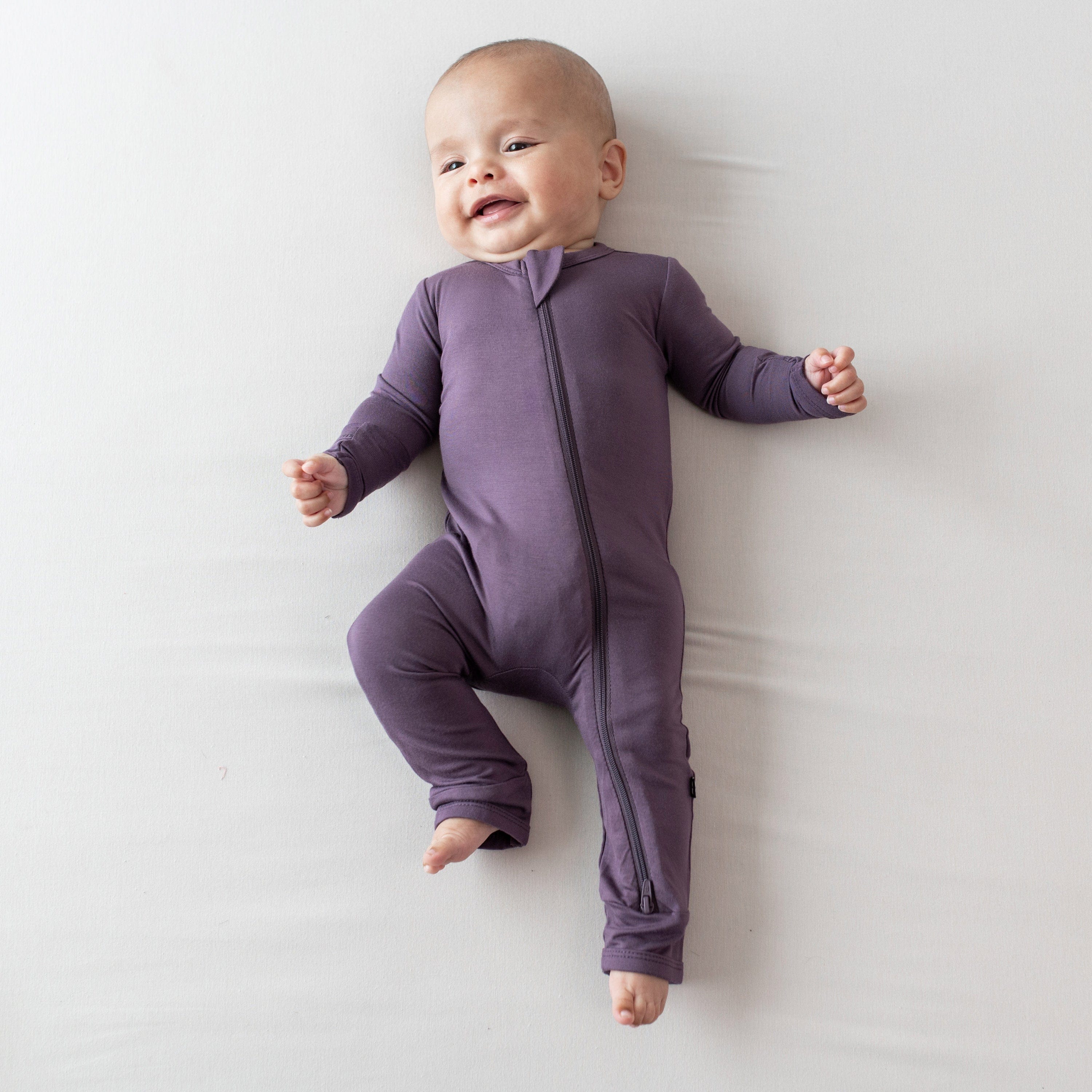 Smiling infant laying on a an off-white surface wearing the Zippered Romper in Currant