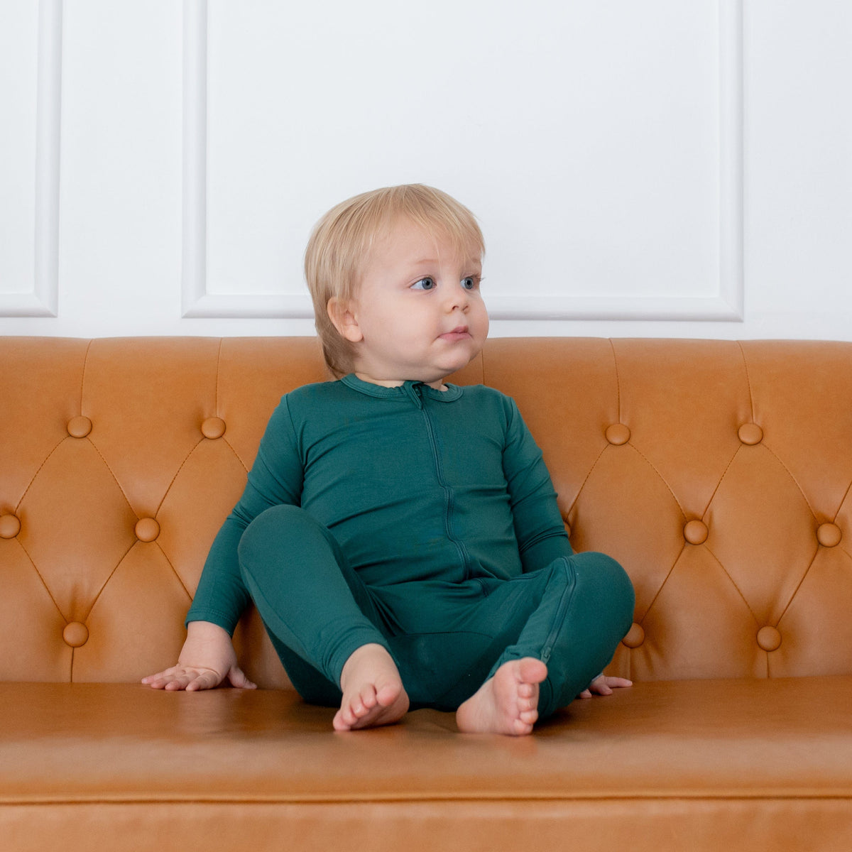 Toddler sitting on a camel leather couch while wearing a Kyte Baby Zippered Romper in Emerald