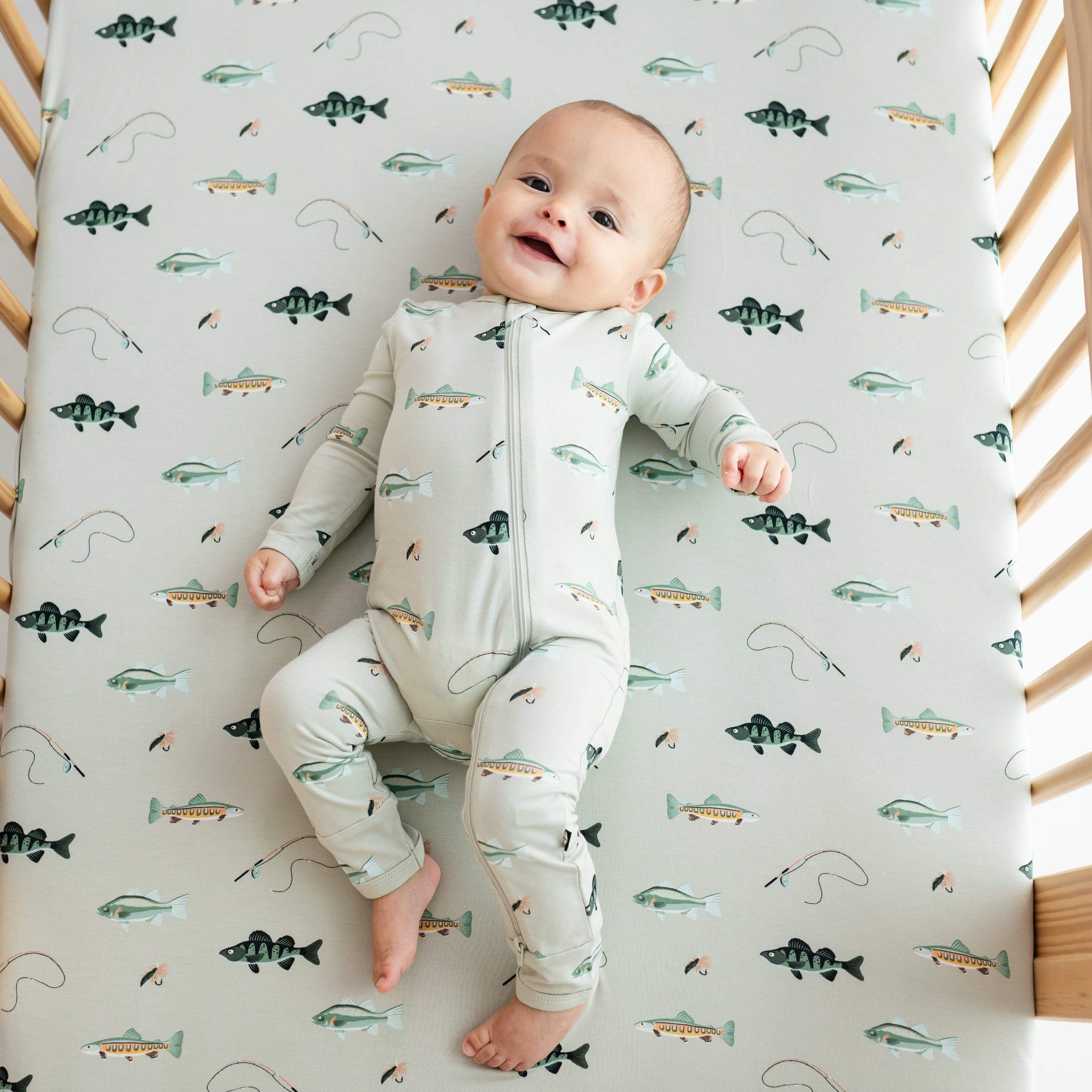 Infant model wearing the Zippered Romper in Fishing laying in a crib on a Fishing Crib Sheet