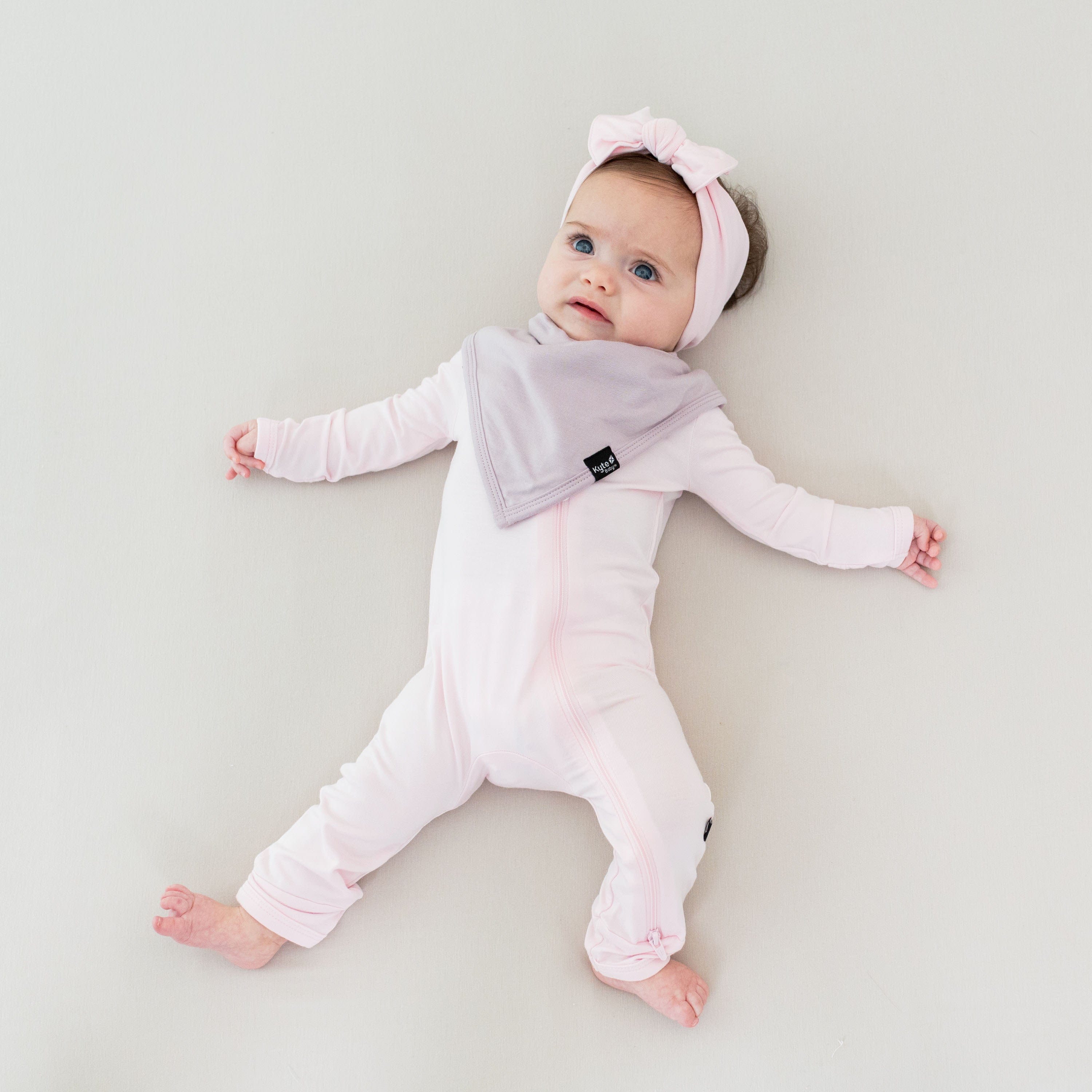 Infant wearing Kyte Baby Bamboo Zippered Romper in Sakura pink with a matching knotted bow headband