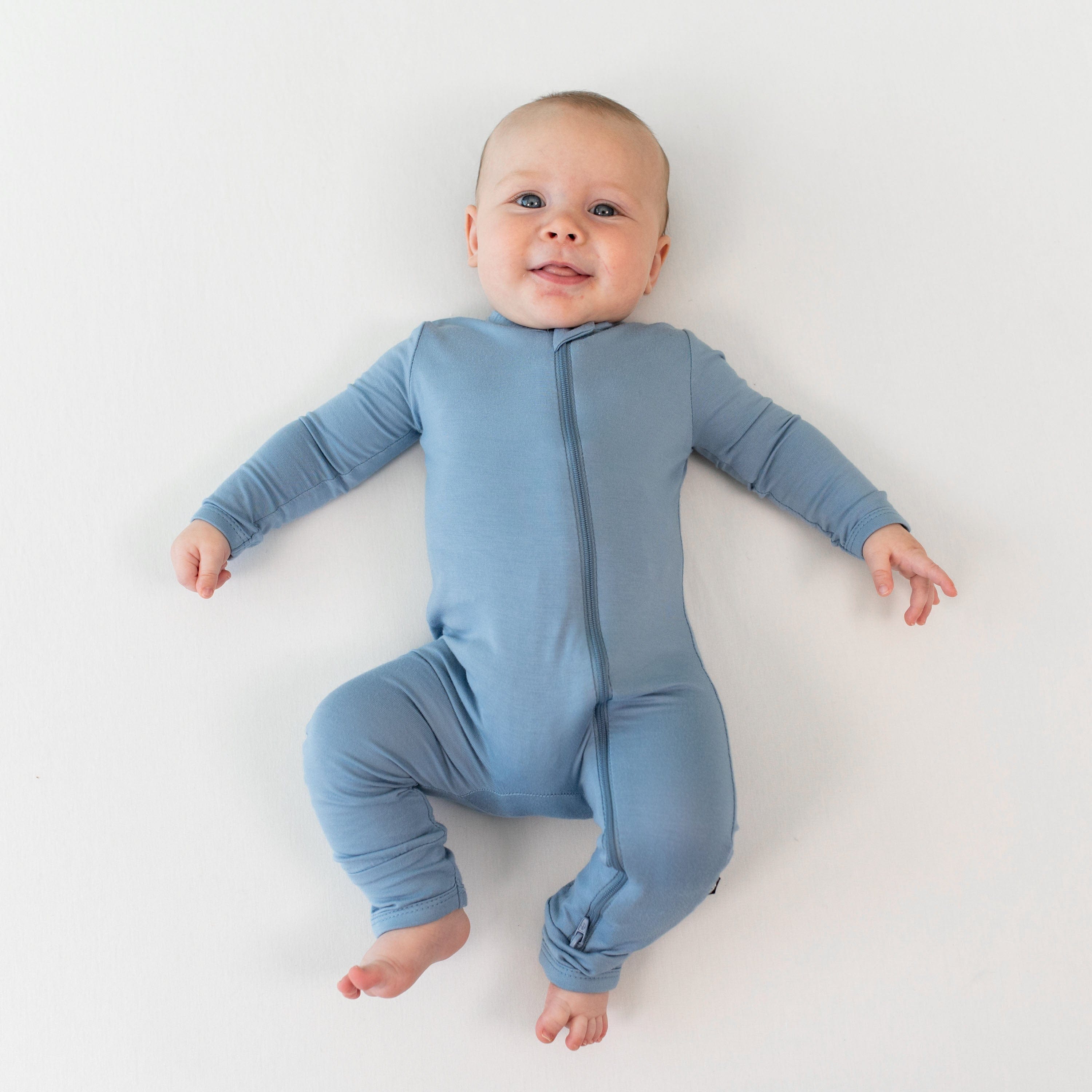 Infant laying on their back on an off-white surface while wearing a Kyte Baby Zippered Romper in Slate