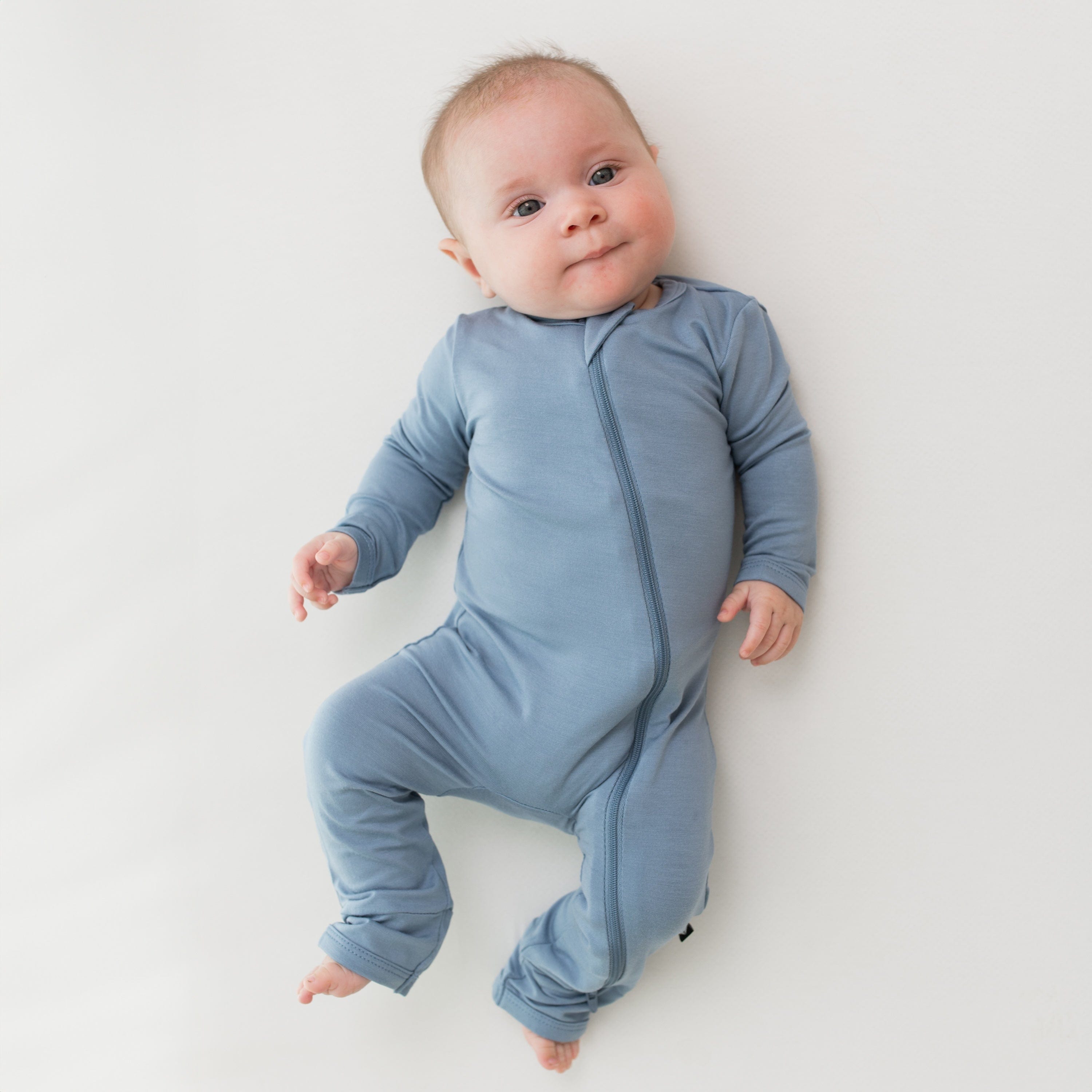 Baby modeling the Kyte Baby Zippered Romper in Slate while laying on a white surface