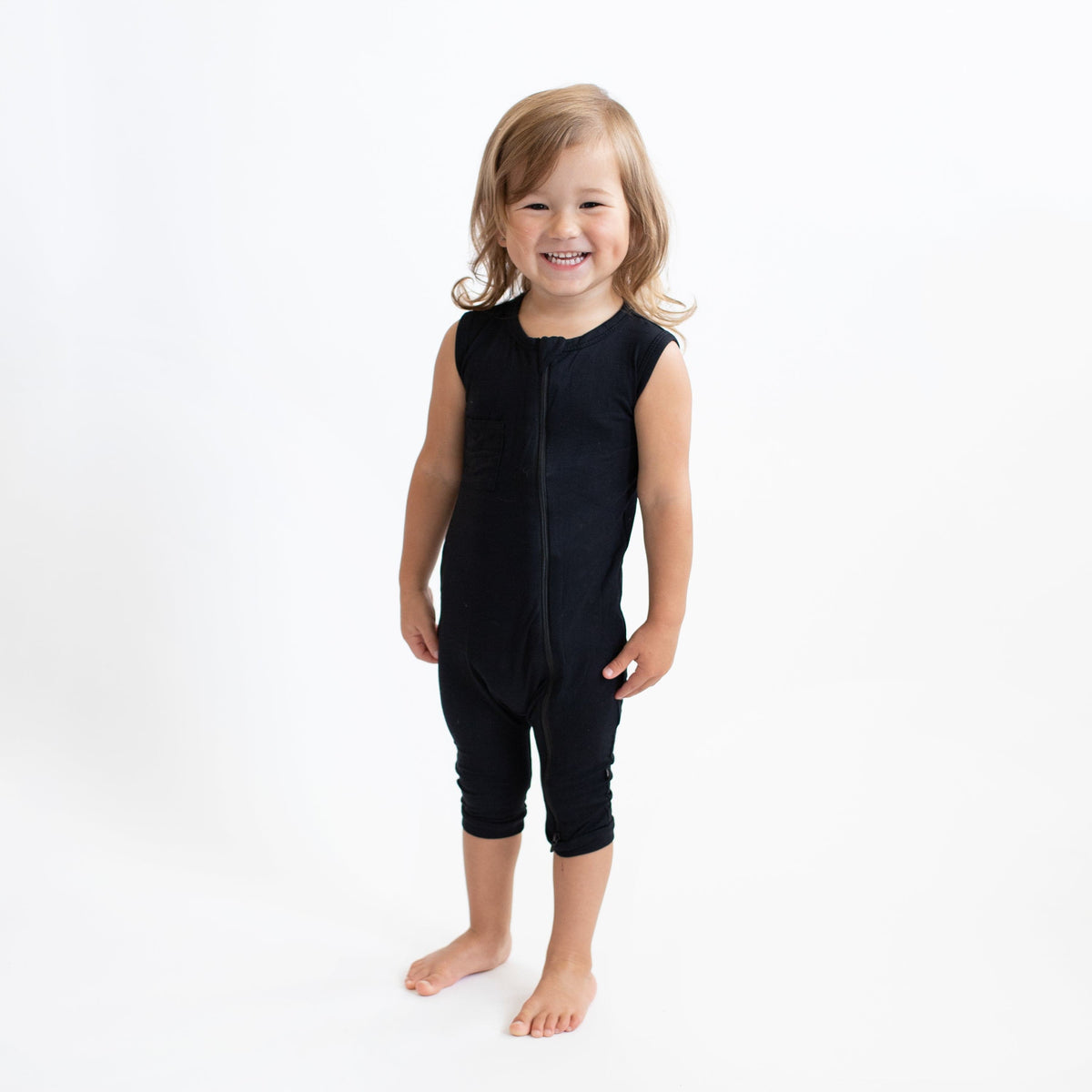 Smiling toddler standing in a white room while wearing Kyte Baby Zippered Sleeveless Romper in Midnight