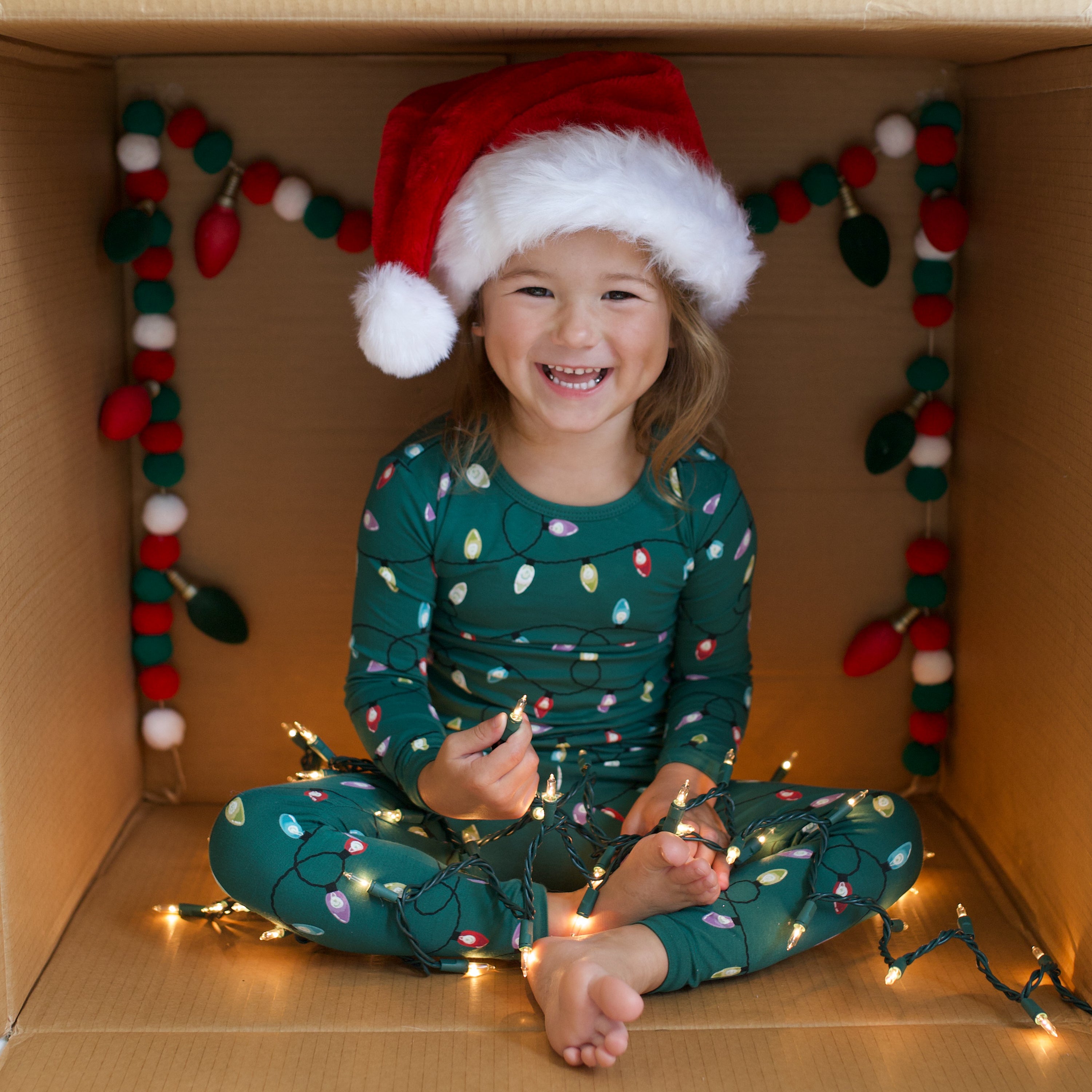 Smiling young girl sitting in a box holding tree lights wearing the Long Sleeve Pajamas in Merry and Bright and a santa hat