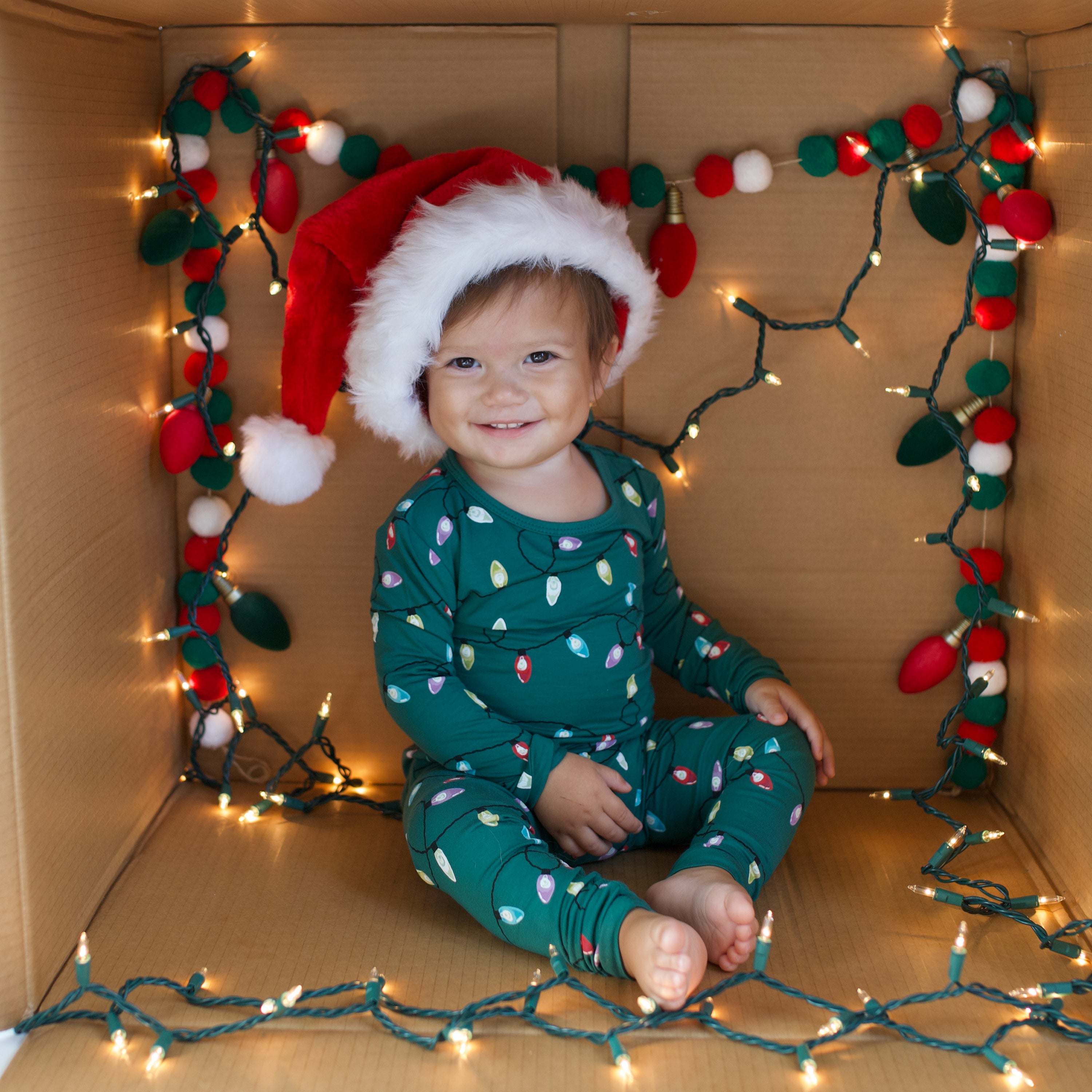 Young toddler sitting in a box wearing the Long Sleeve Pajamas in Merry and Bright surrounded by Christmas lights