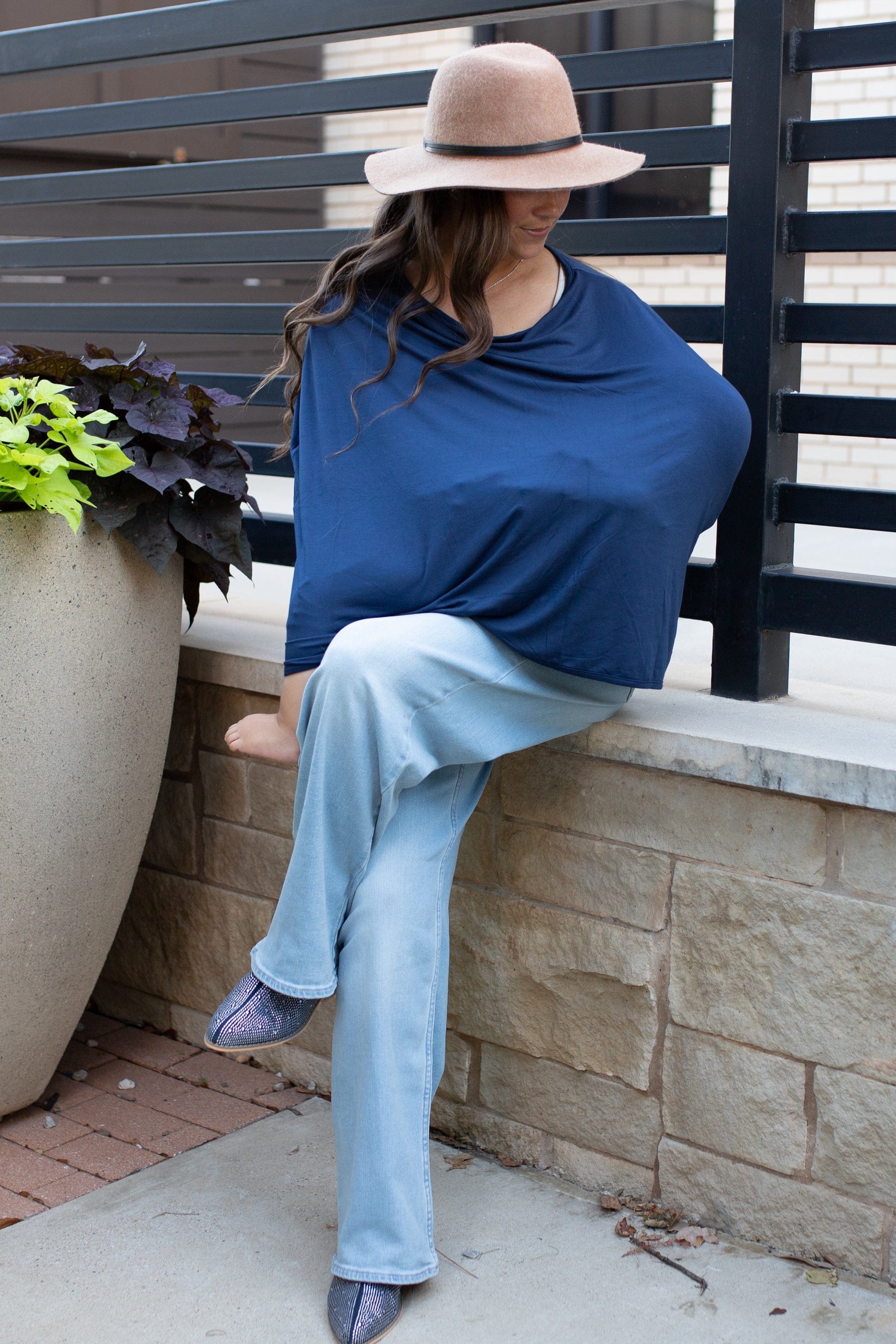 Woman sitting on a ledge while nursing a baby with a navy blue nursing cover on with a potted plant nearby.