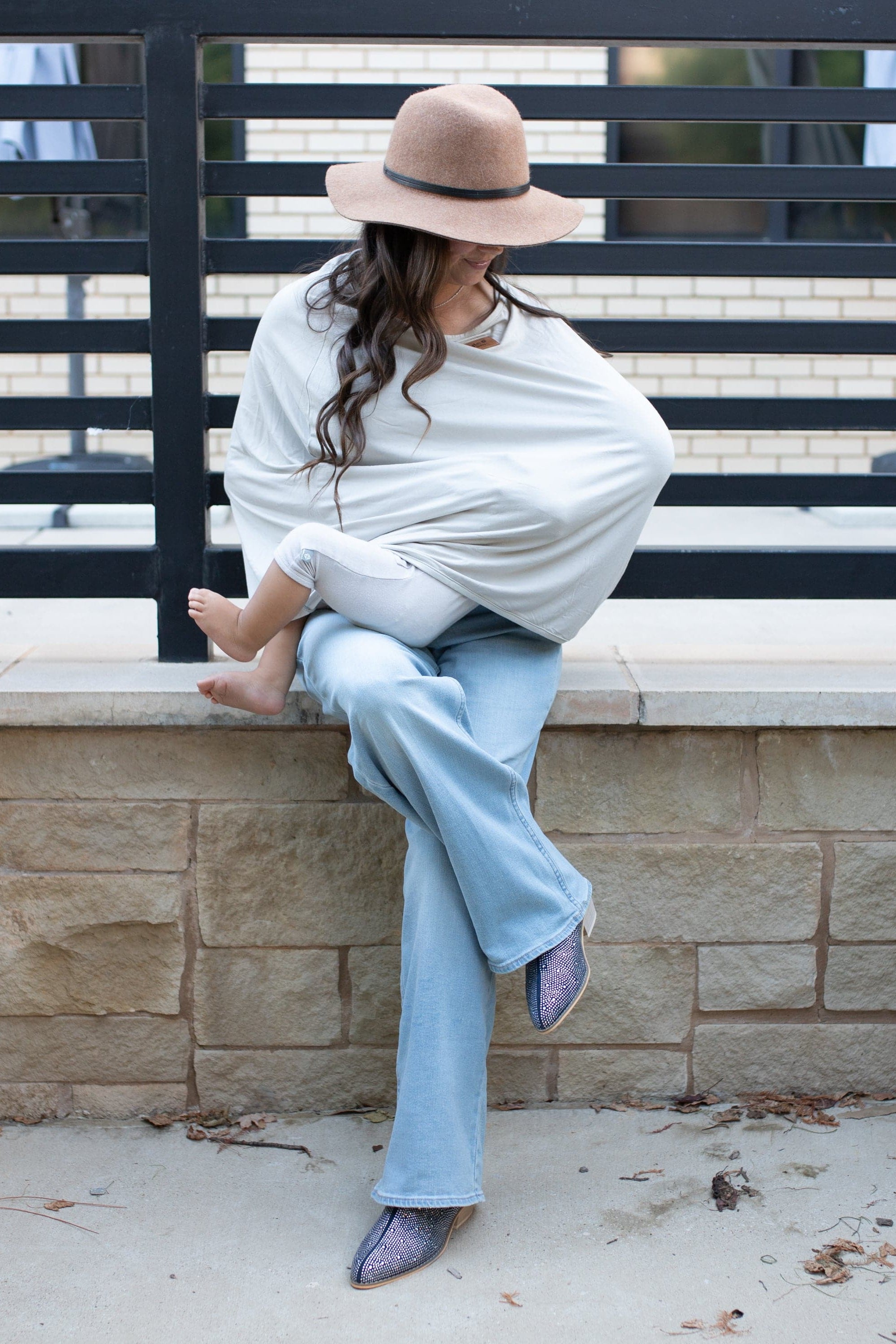 Woman sitting on a stone ledge with a baby nursing, wearing a nursing cover, wide-brimmed hat and light blue pants.