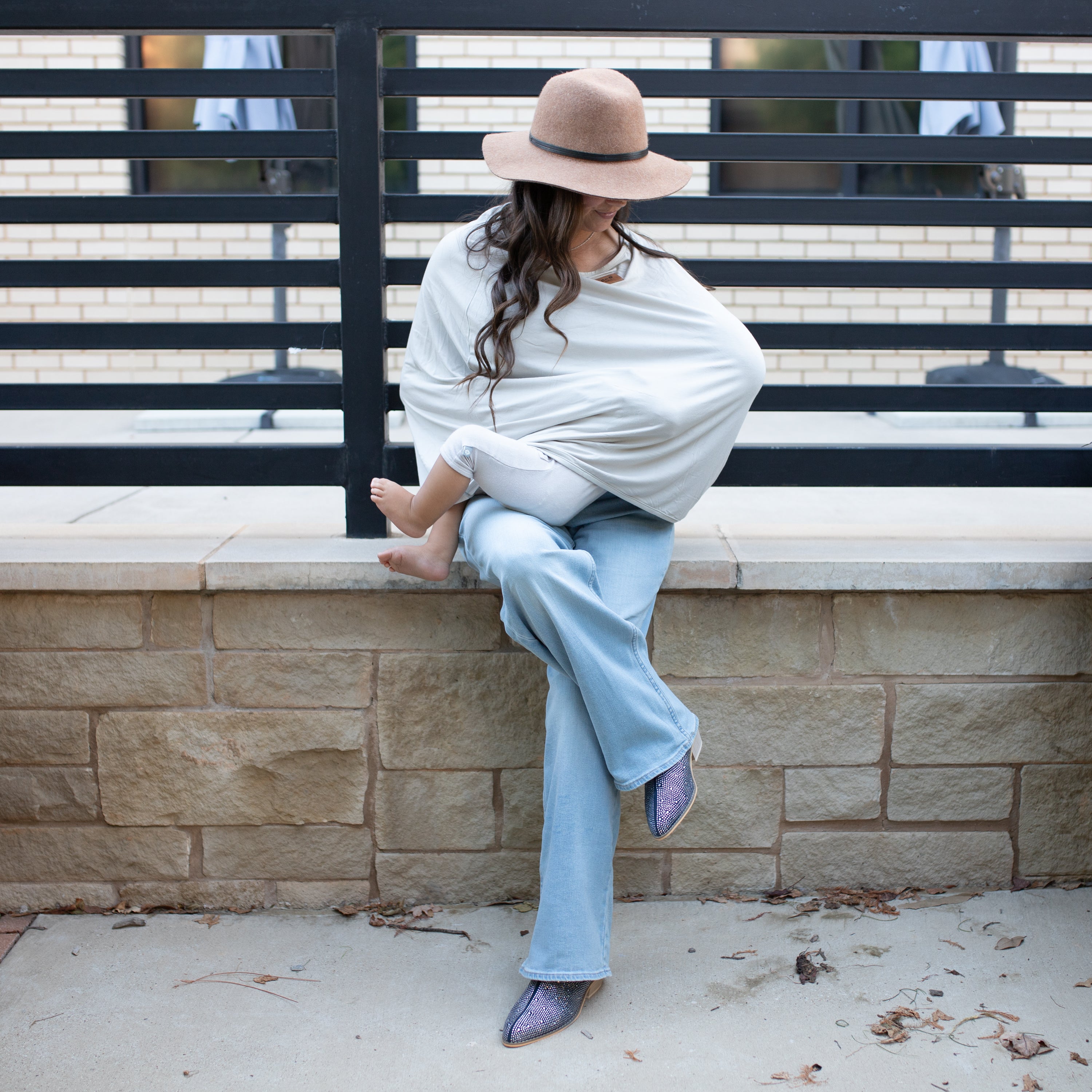 Woman sitting on a stone ledge with a baby nursing, wearing a nursing cover, wide-brimmed hat and light blue pants.