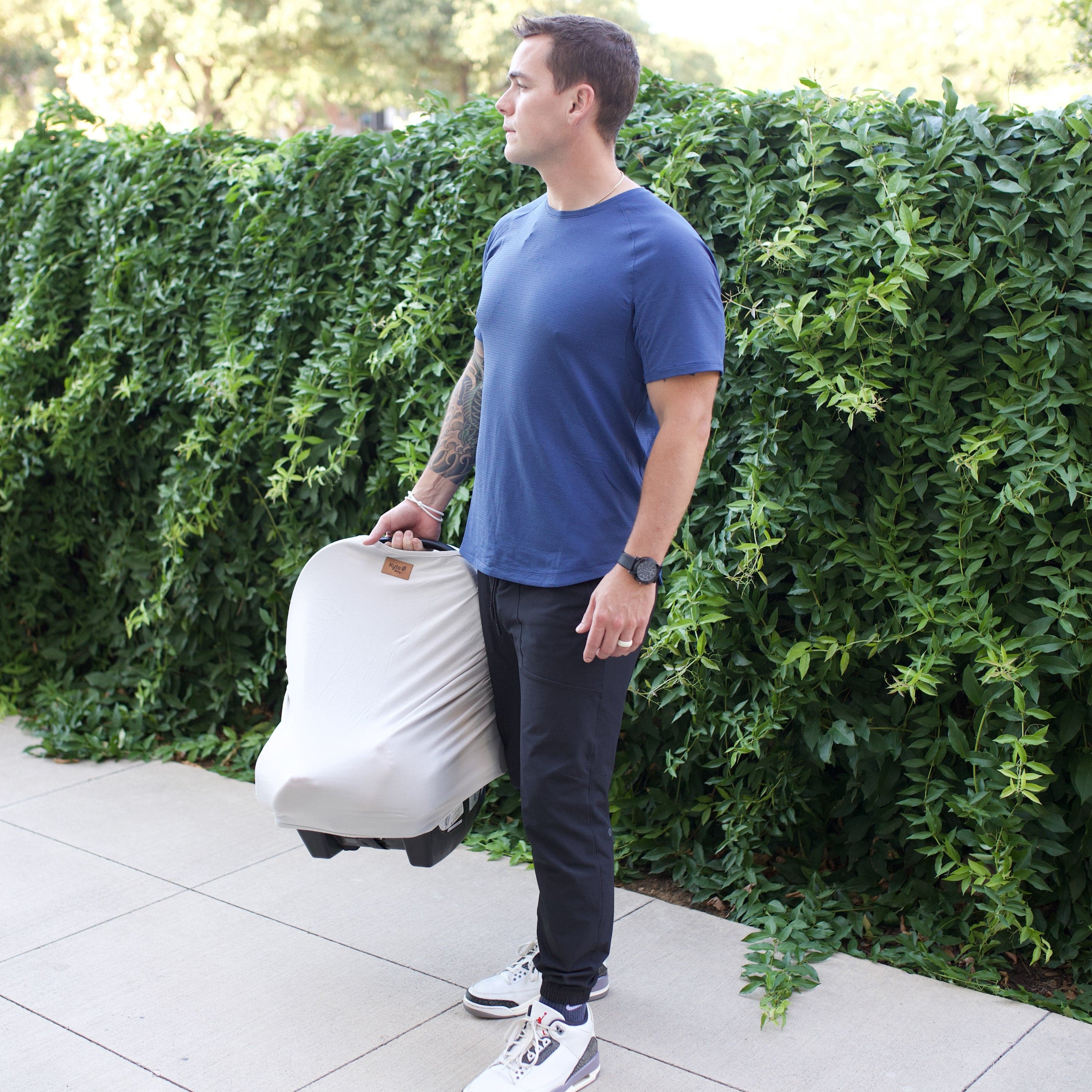 Man holding a car seat that has a light grey car seat cover on it while outdoors with greenery in the background
