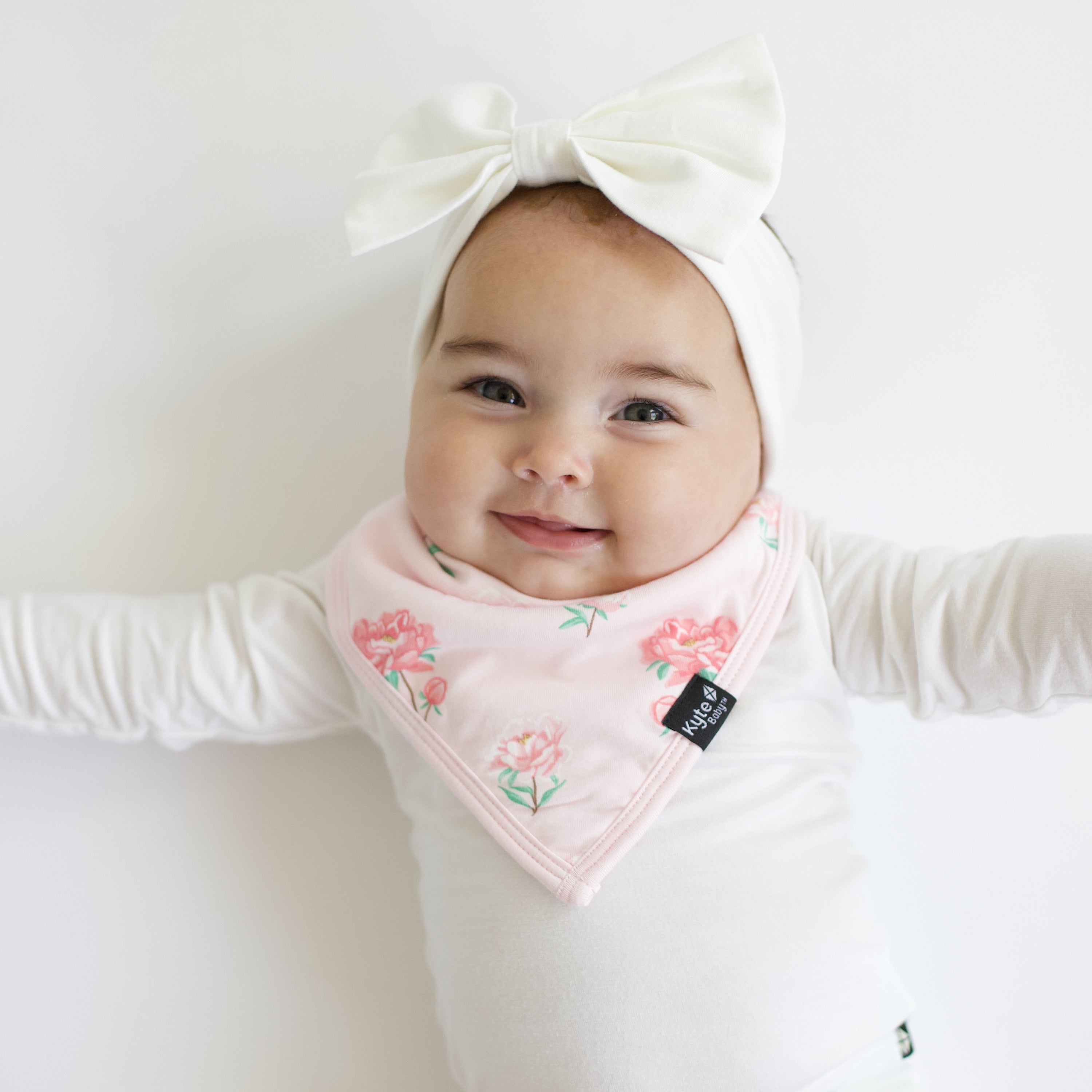Smiling baby wearing the Bib in Sakura Peony with a long sleeve Cloud bodysuit and matching Bow Headband