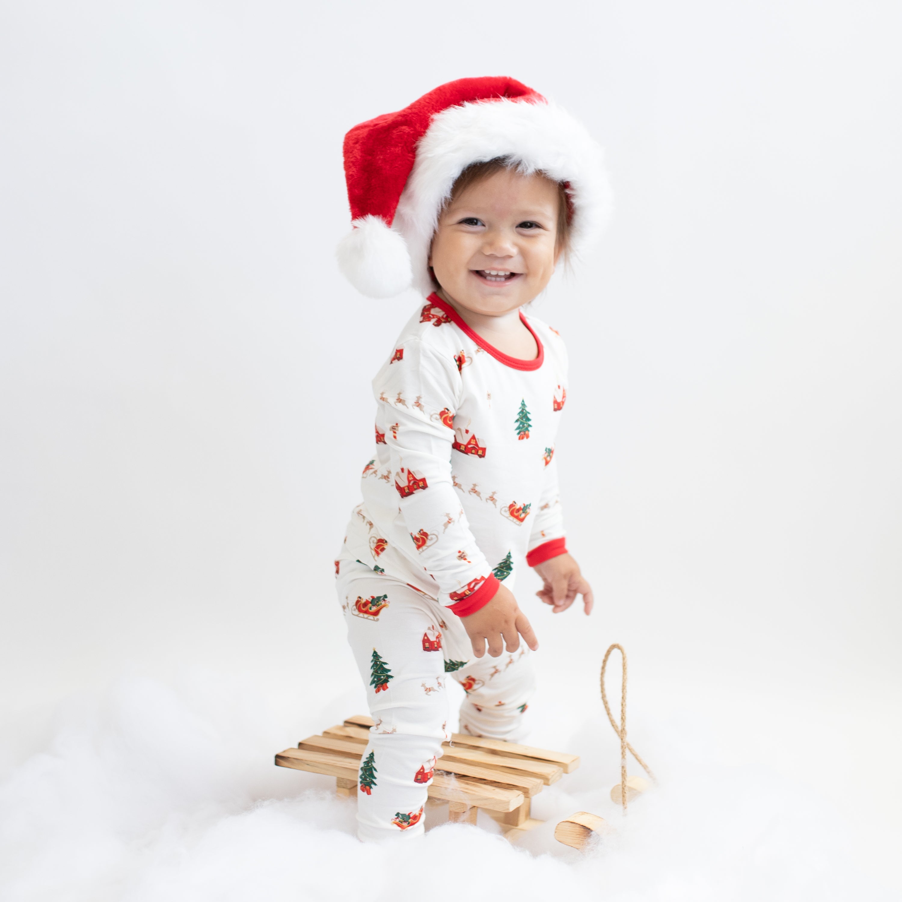 Young smiling toddler standing over a toy wooden sleigh wearing the Long Sleeve Pajamas in Santa Sleigh with a red and white santa hat