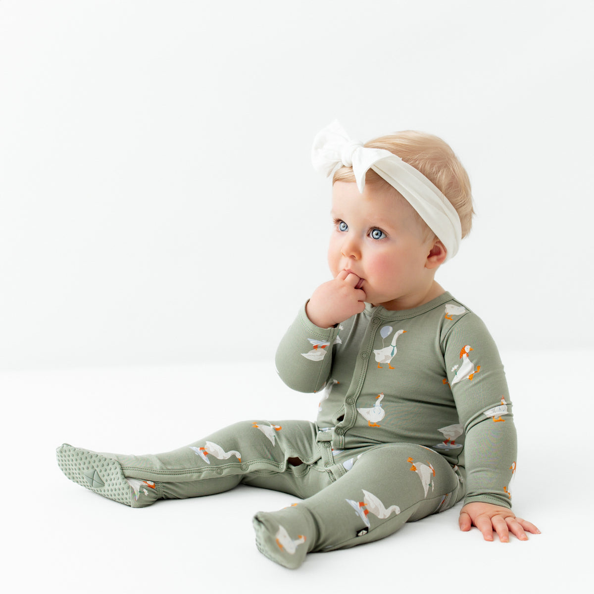 Infant sitting with fingers in her mouth wearing the Footie in Silly Goose paired with a Cloud Knotted Bow Headband