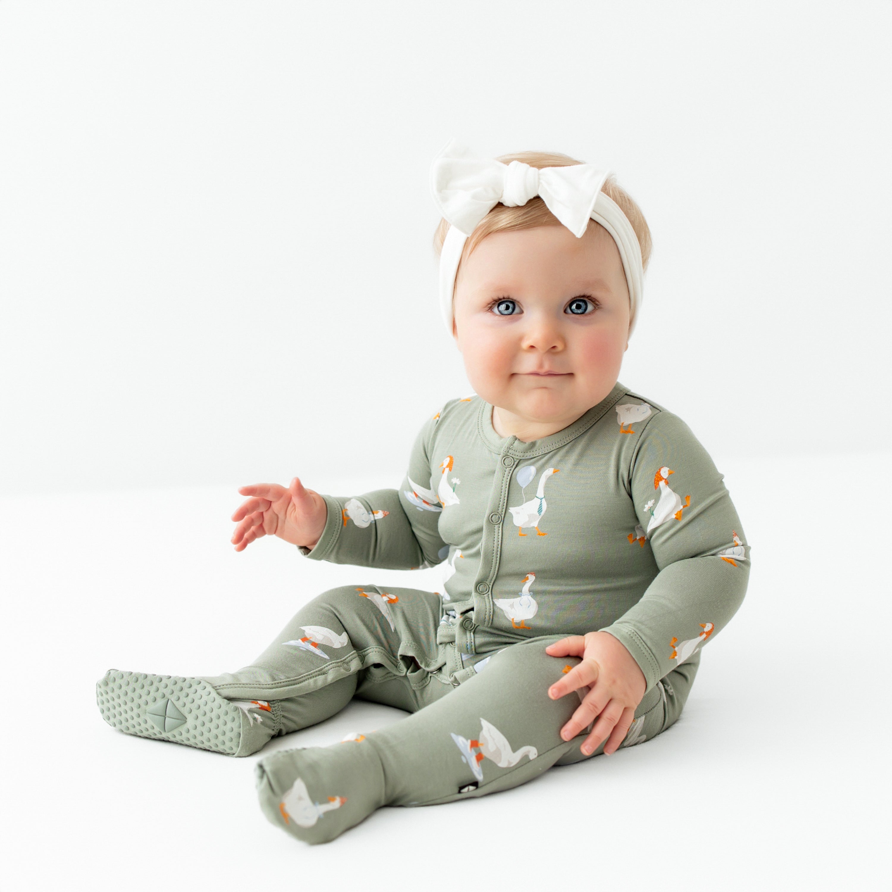 Smiling infant sitting wearing the Footie in Silly Goose paired with a Cloud Knotted Bow Headband