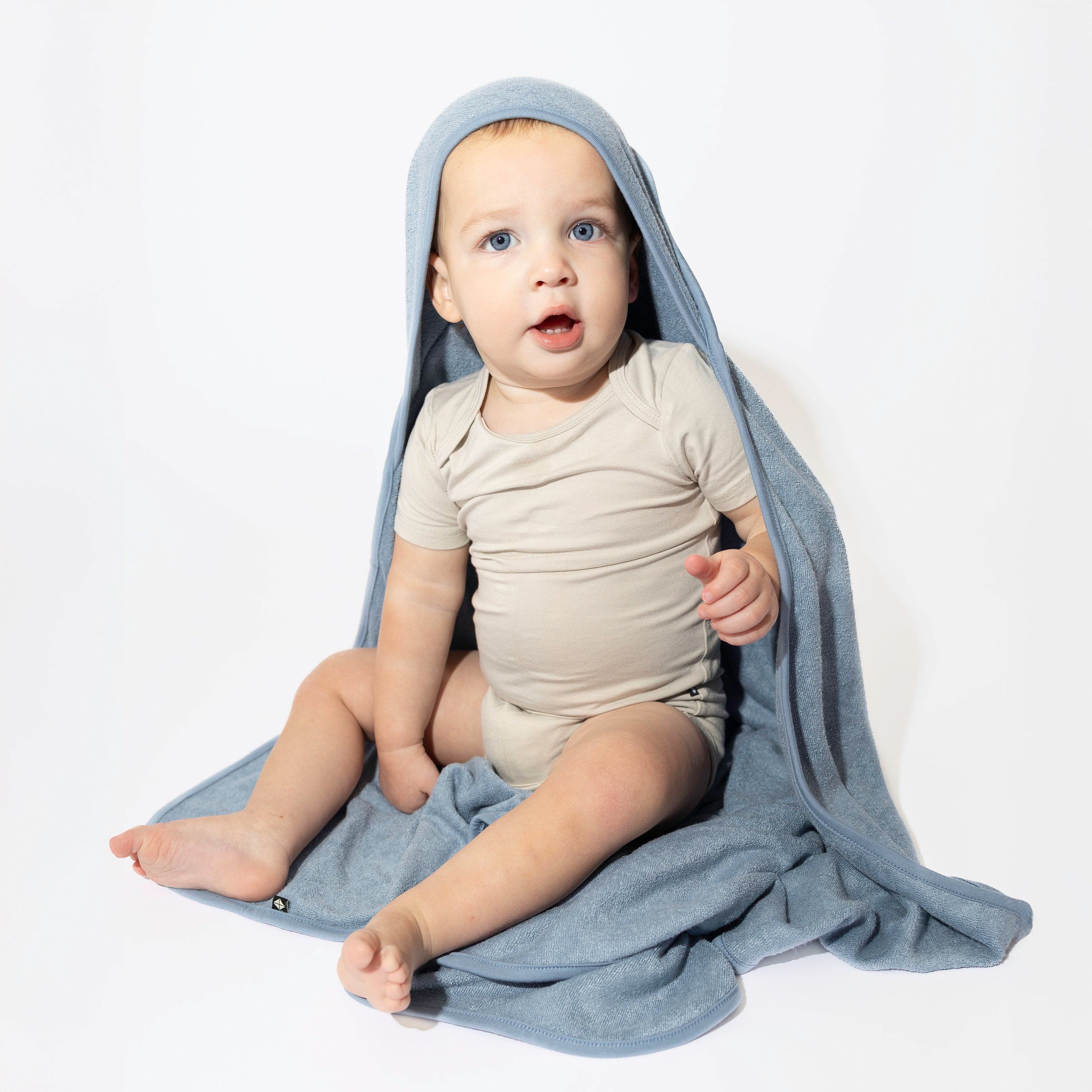 Young toddler sitting on a white surface wearing the Hooded Bath Towel in Slate