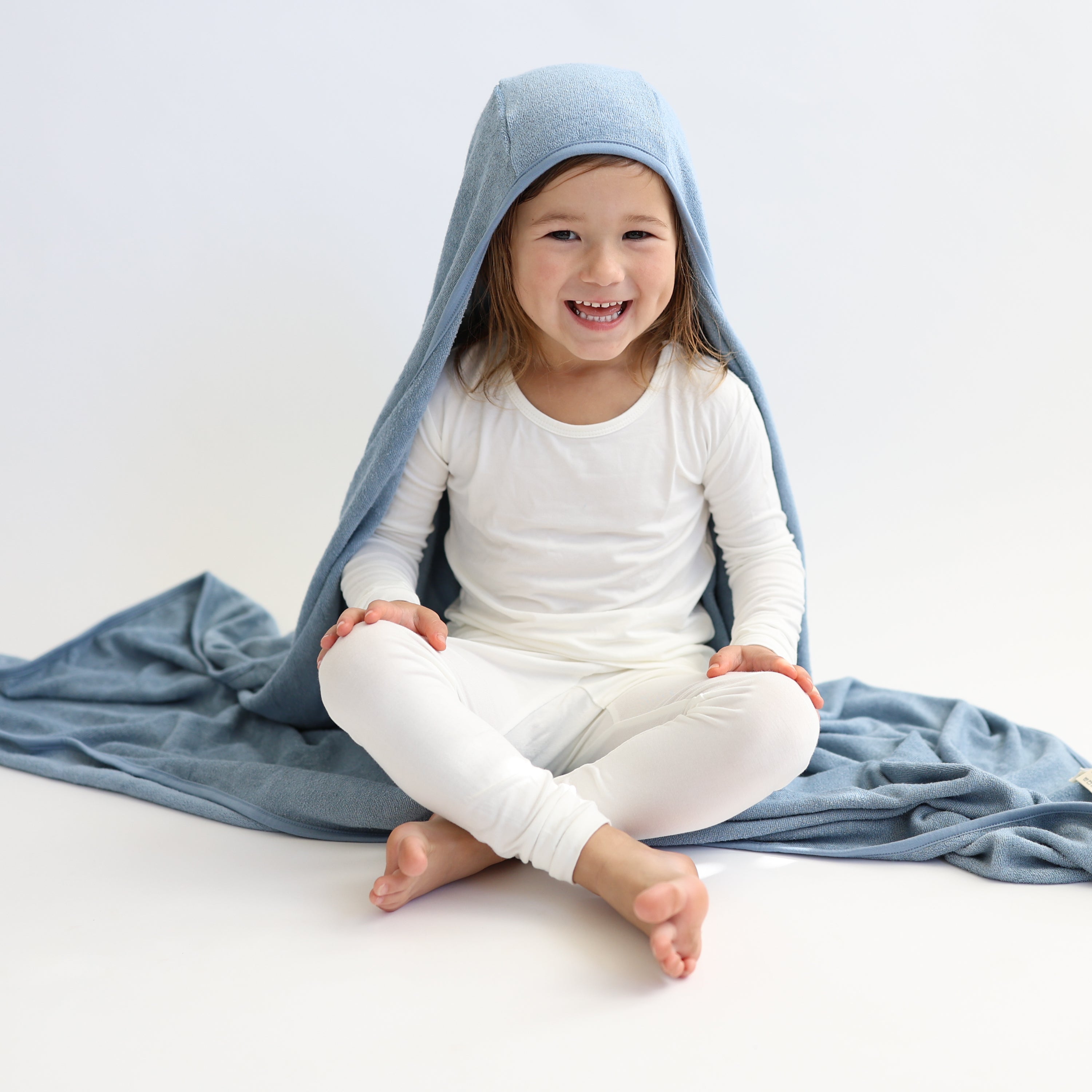 Young smiling girl sitting on a white surface wearing the Toddler Hooded Bath Towel in Slate