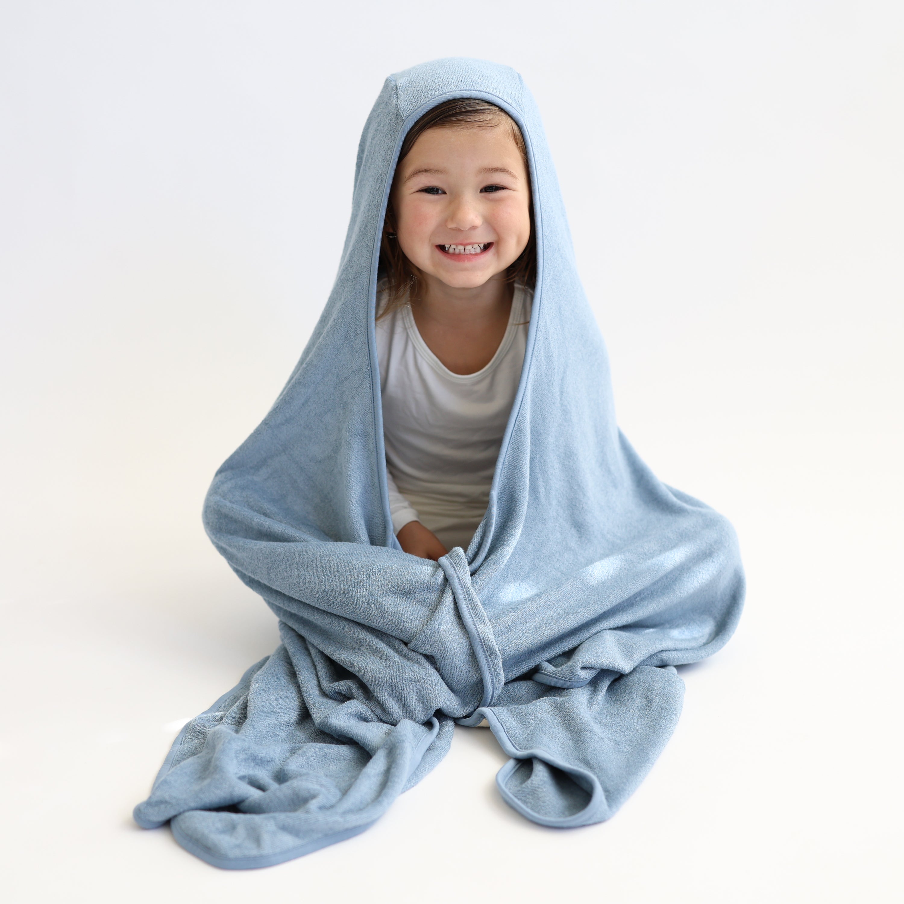 Young girl sitting on a white surface wrapped in the Toddler Hooded Bath Towel in Slate