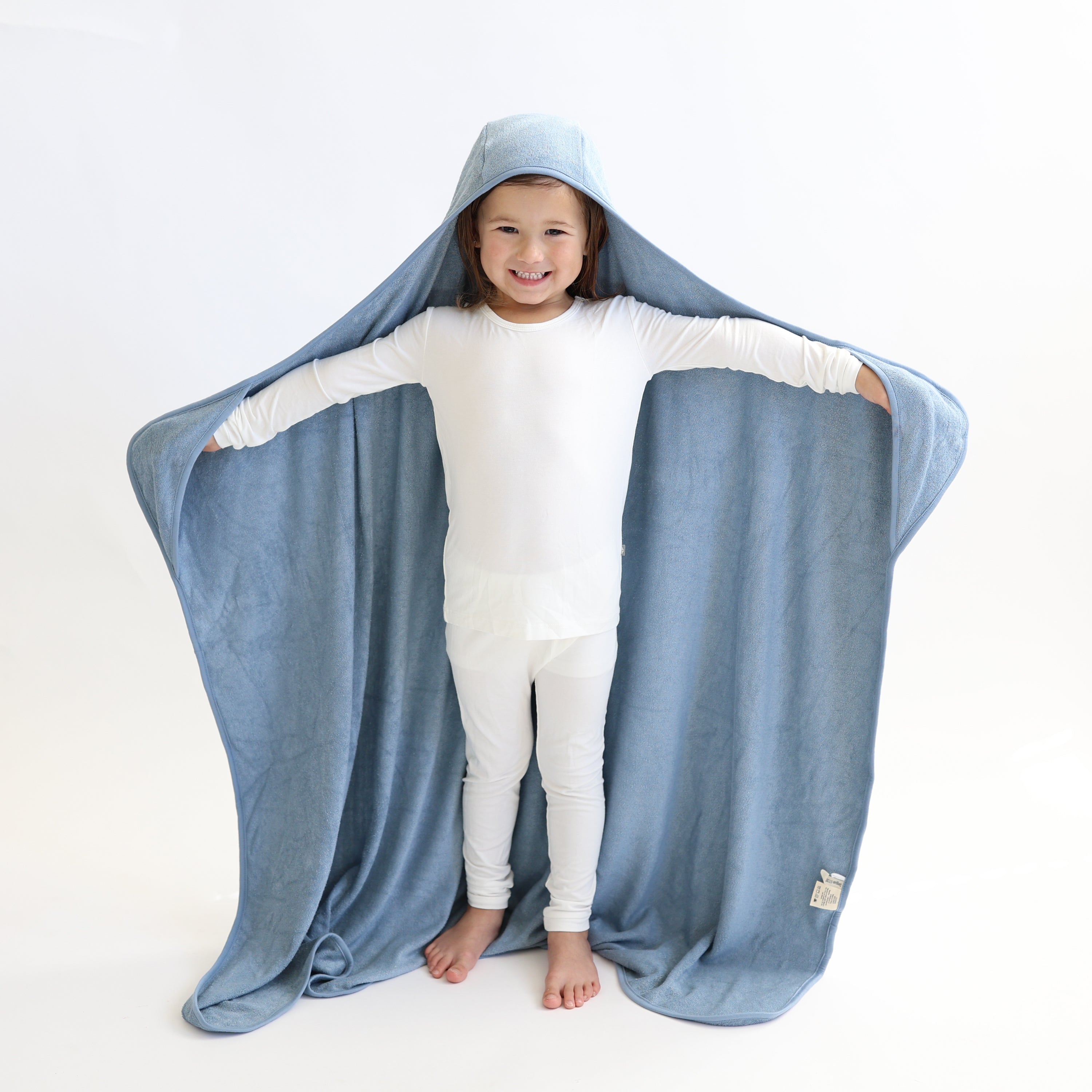 Young girl standing wearing the Toddler Hooded Bath Towel in Slate with her head in the hood and hands in the pockets of the towel