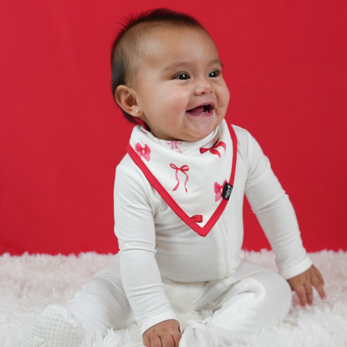 Infant sitting on a white plush carpet in front of a red background wearing a white footie with the absorbent Bib in Small Love Bow