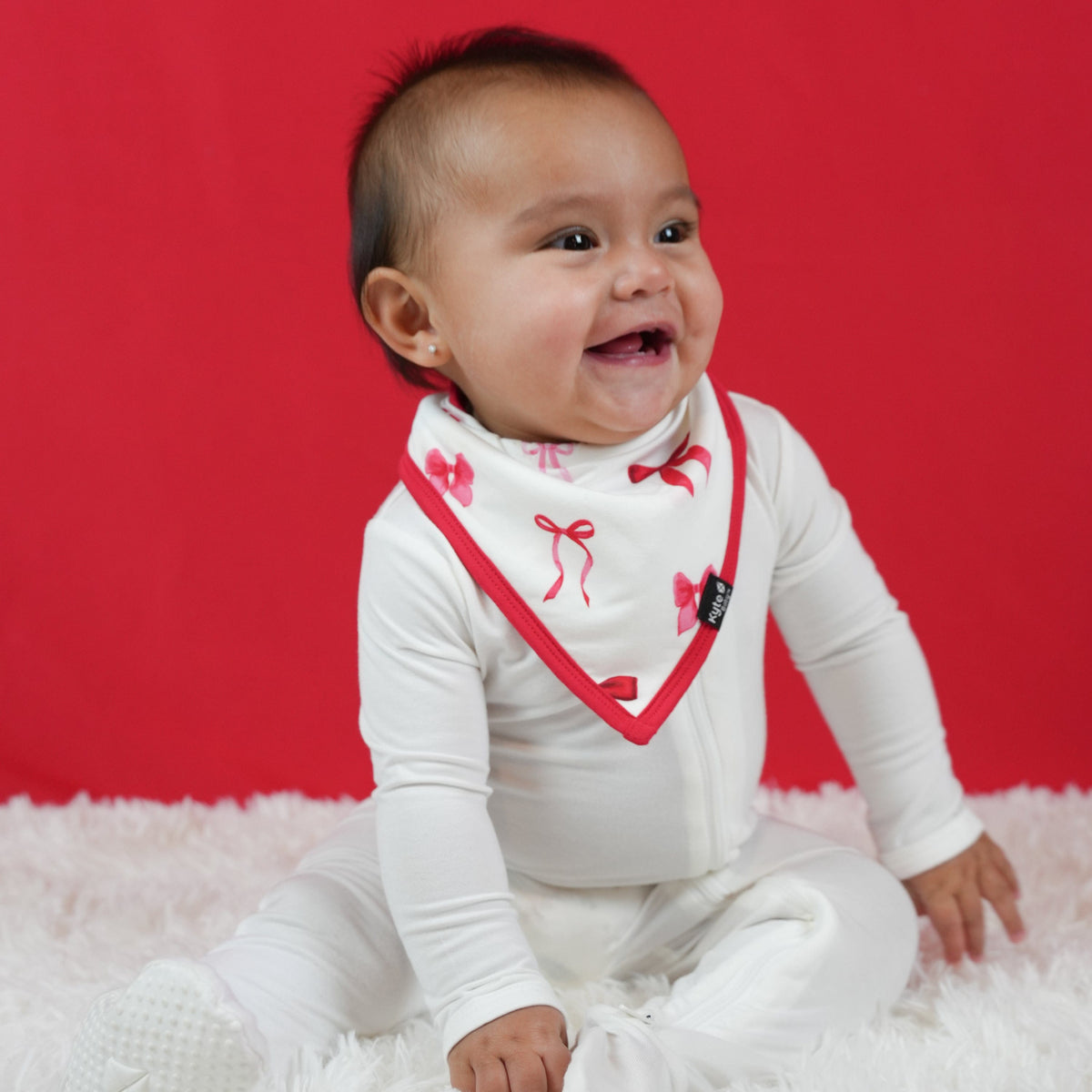 Infant sitting on a white plush carpet in front of a red background wearing a white footie with the absorbent Bib in Small Love Bow