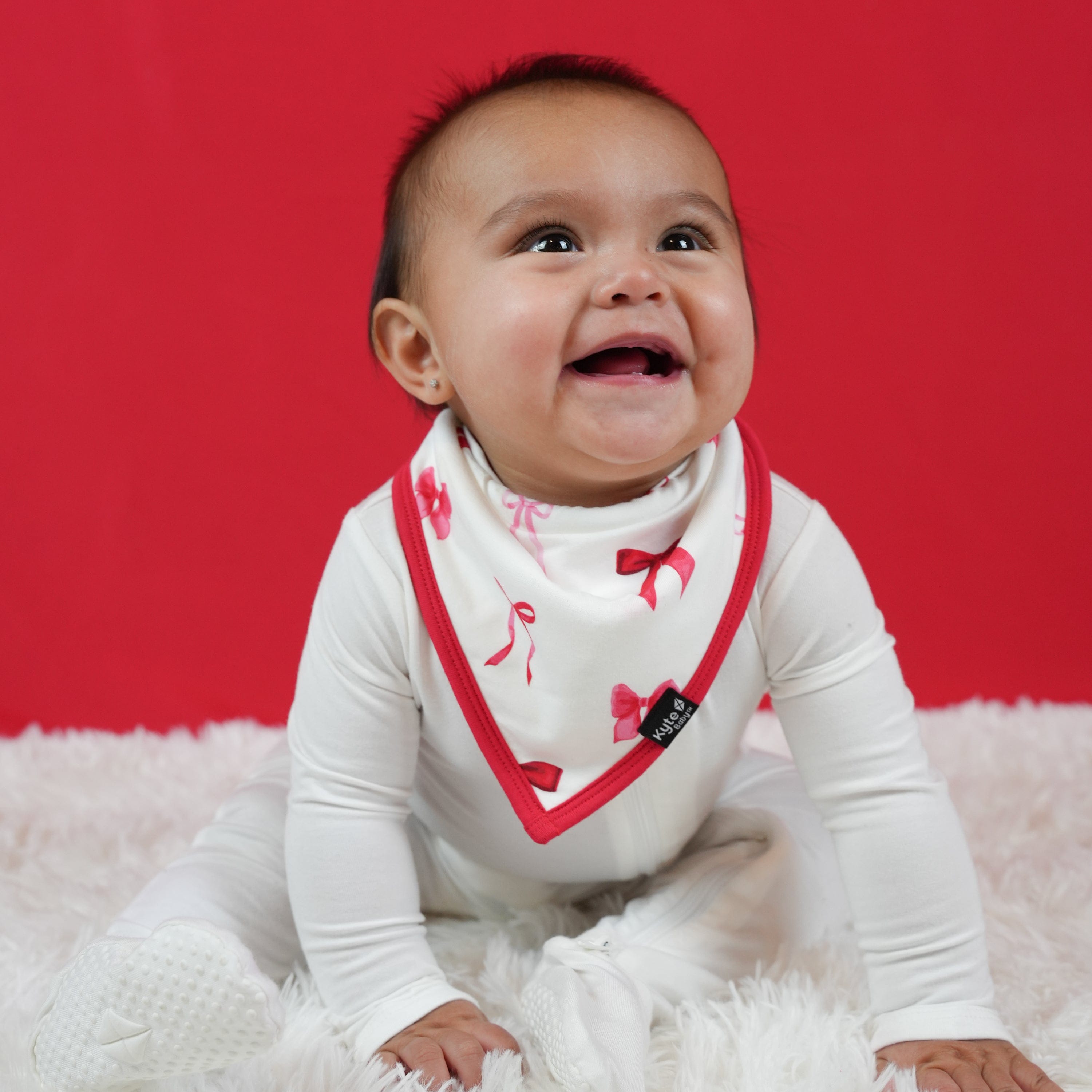 Smiling infant sitting on a white plush carpet in front of a red background wearing a white footie with a Bib in Small Love Bow