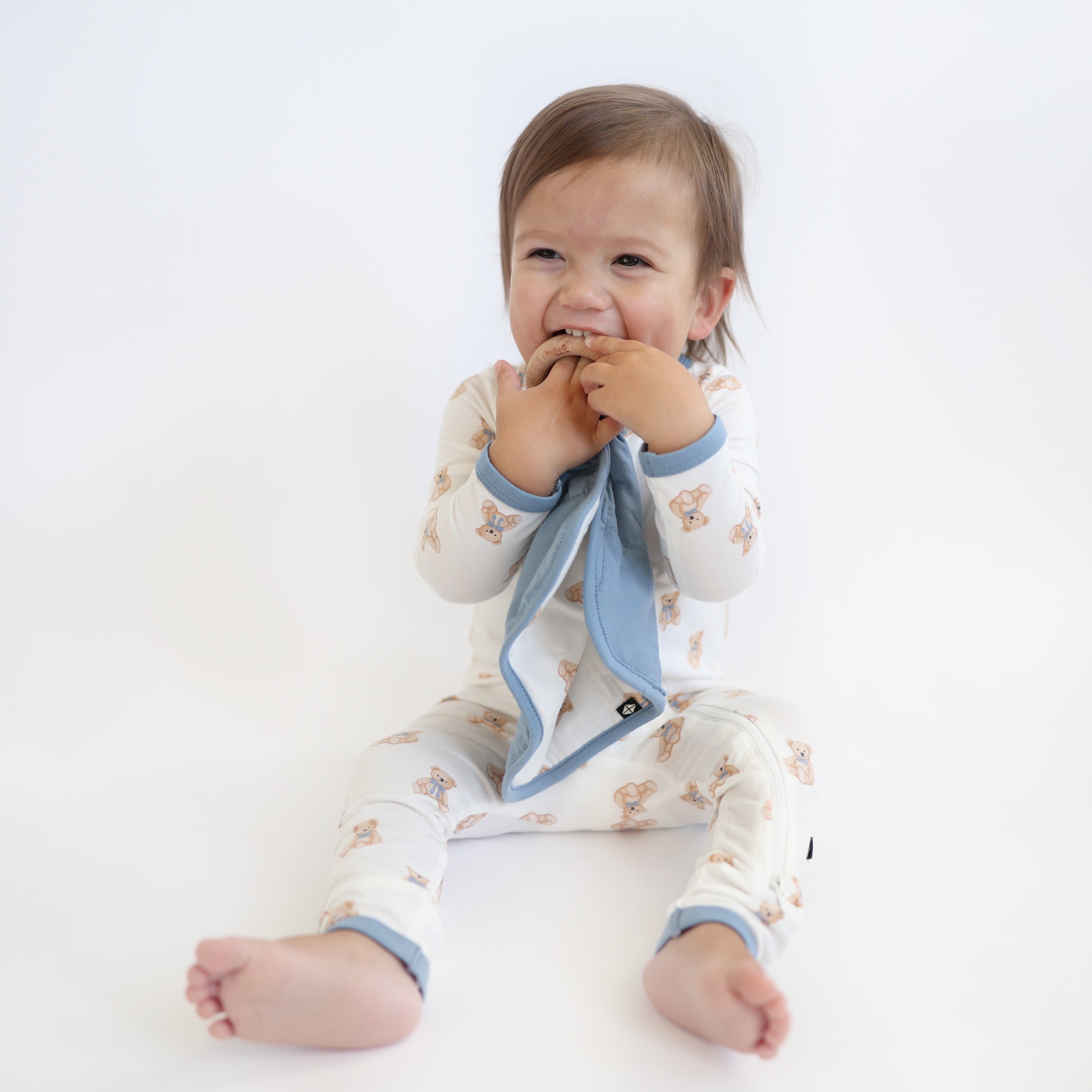 Young toddler sitting on a light neutral surface holding the Lovey in Teddy Bear with Removable Wooden Teething Ring and chewing on the wooden ring wearing matching zippered romper
