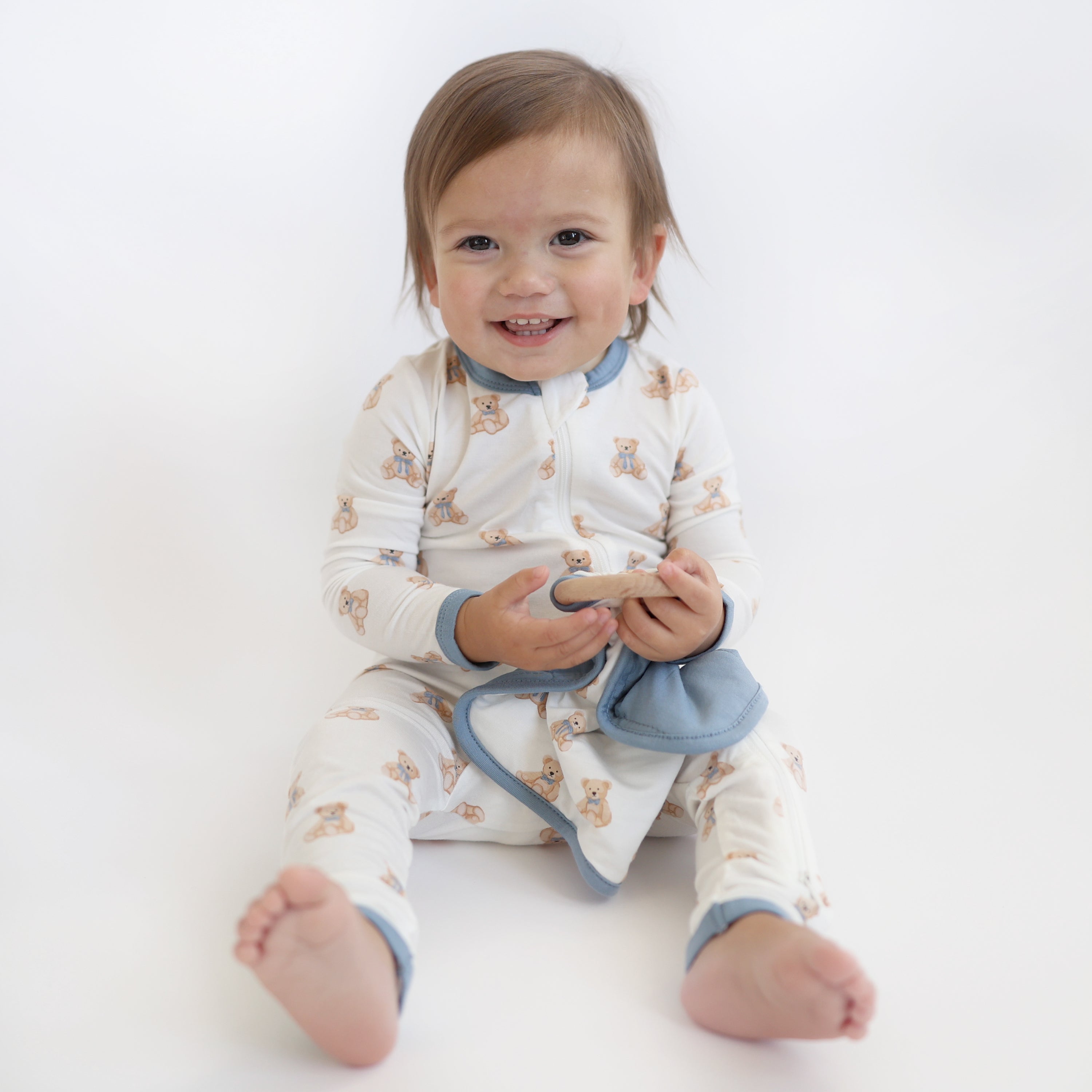 Smiling toddler sitting on the floor holding the Lovey in Teddy Bear with Removable Wooden Teething Ring wearing a matching zippered romper