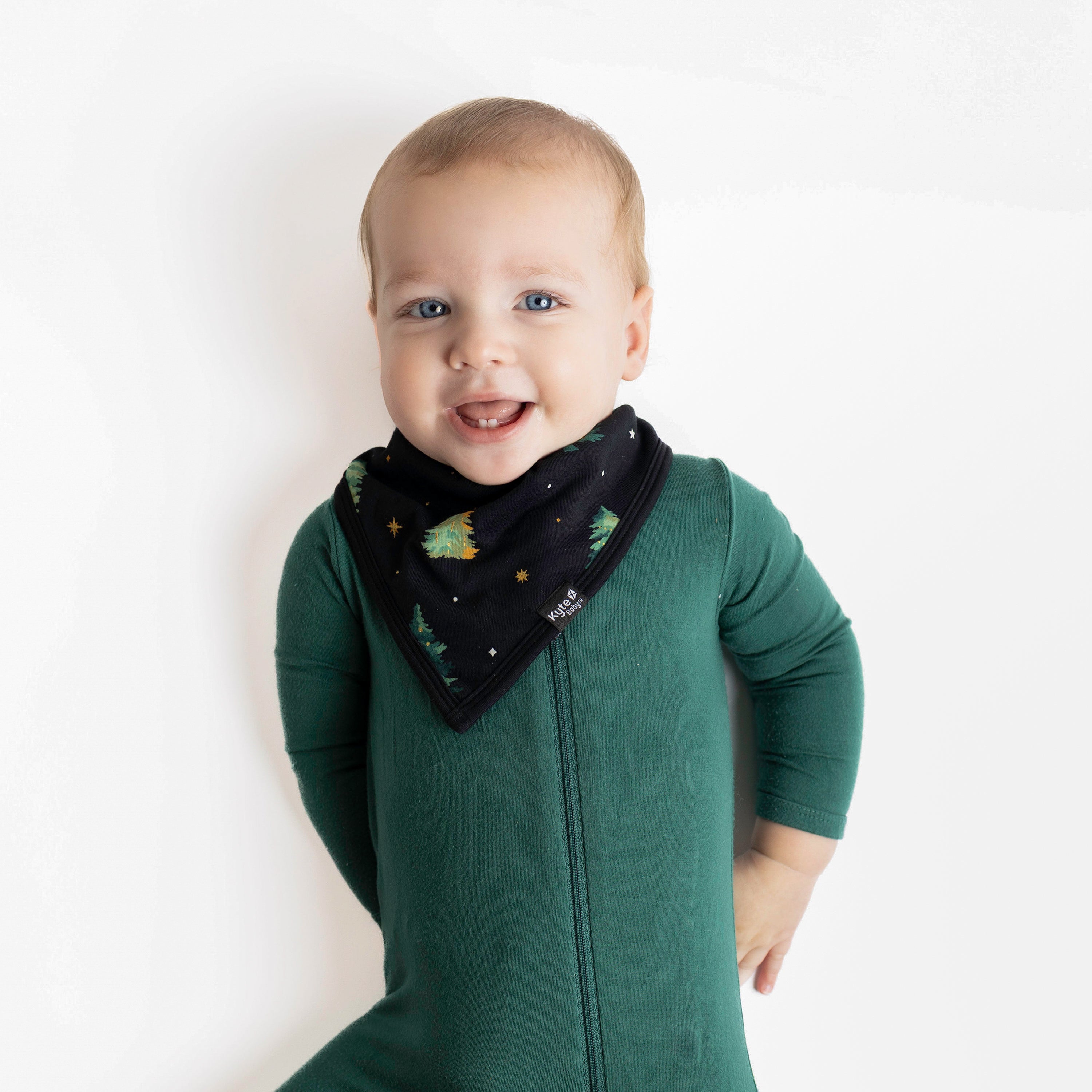 Young toddler laying down wearing the Bib in Twinkle Tree paired with an Emerald Zipper romper