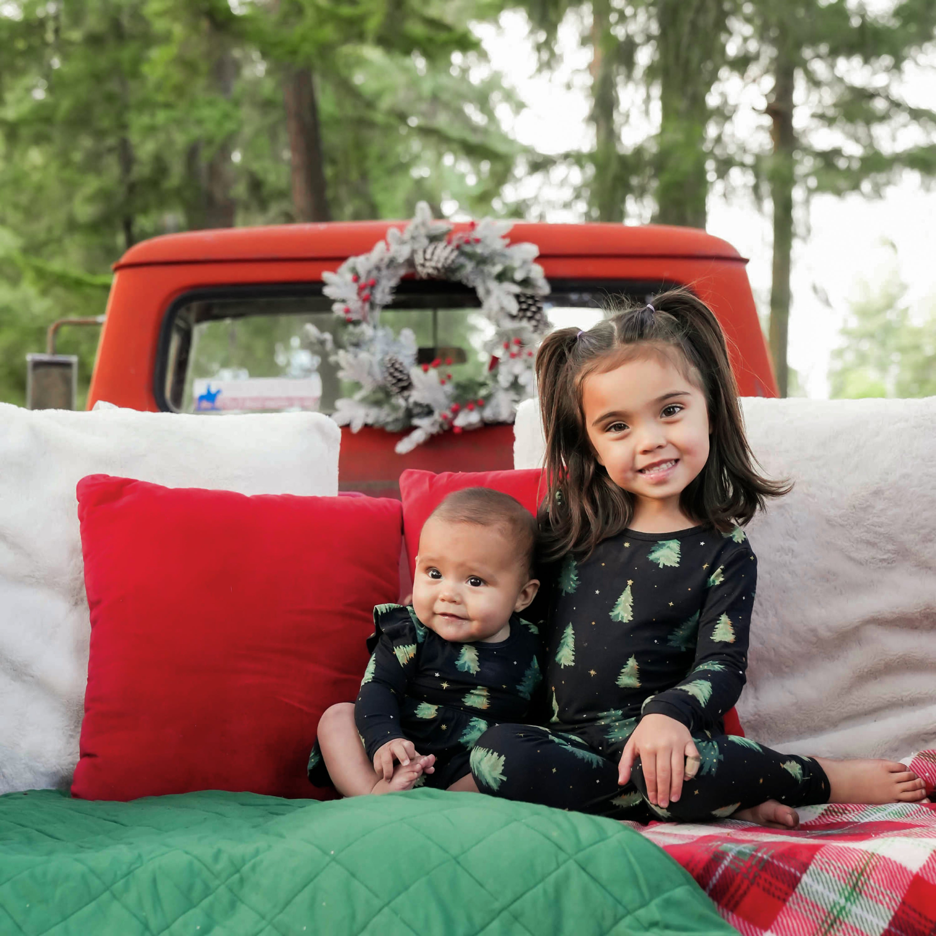 Two sisters sitting on a green and red and white plaid blanket in front of white and red pillows matching in Twinkle Trees in the truck bed of a red truck. Big sister is wearing the long sleeve toddler pajama set and younger sister is wearing the long sleeve twirl bodysuit dress