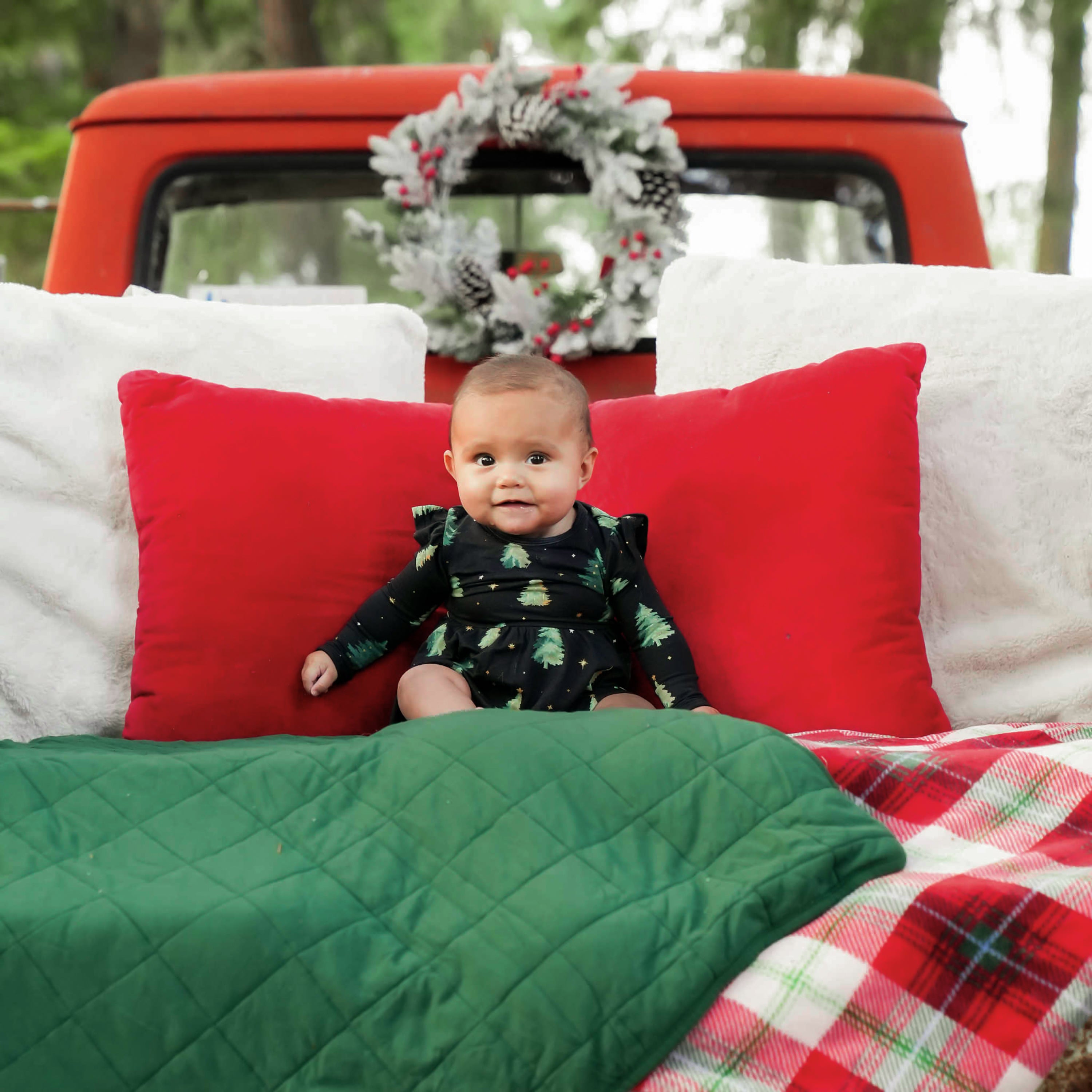 Infant sitting in the back of a truck bed on top of a green and red and white plaid blankets wearing the Long Sleeve Twirl Bodysuit Dress in Twinkle Tree with red and white pillows behind her