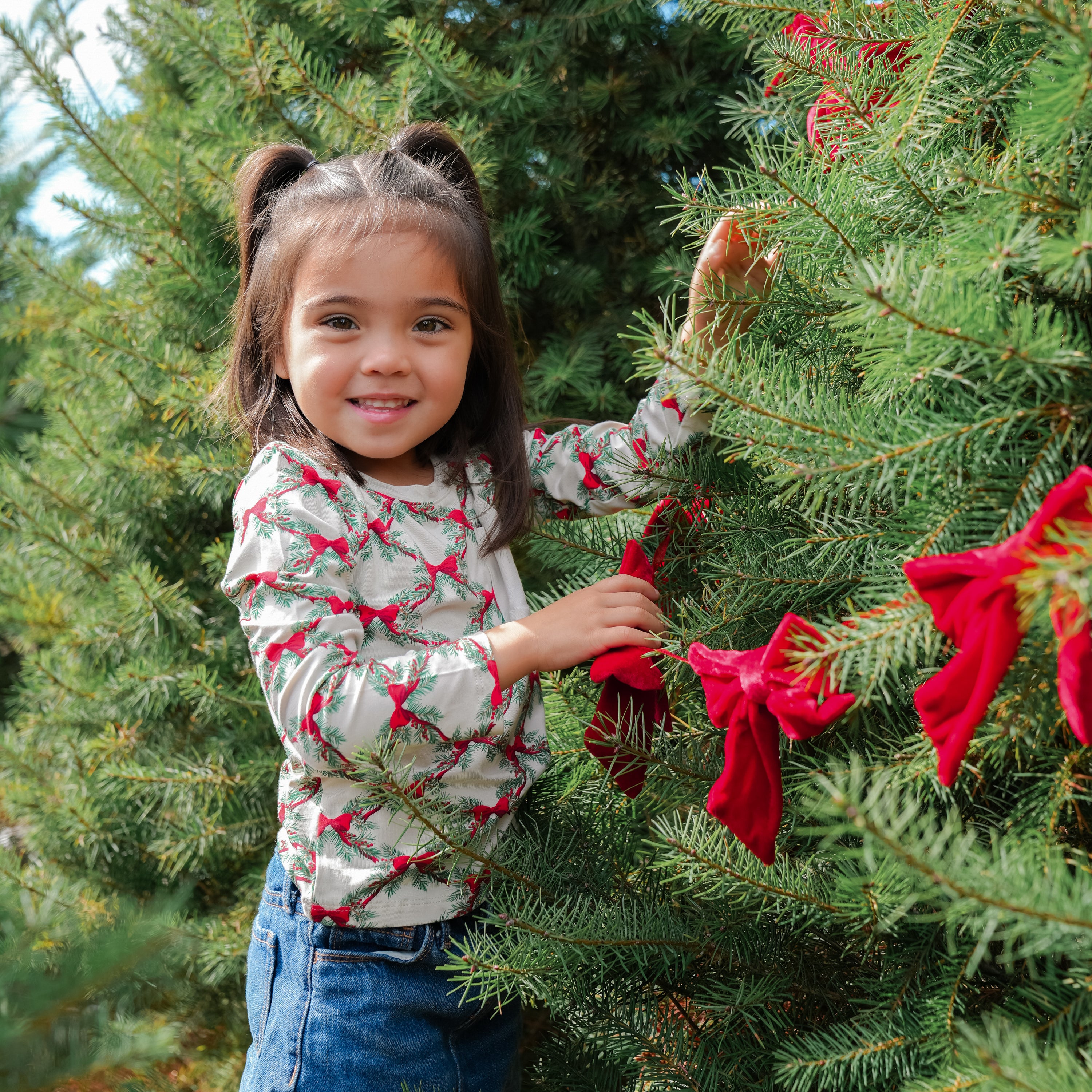 Young girl wearing the Long Sleeve Toddler Crew Neck Tee in Holiday Bow standing beside a green tree decorated with red bows