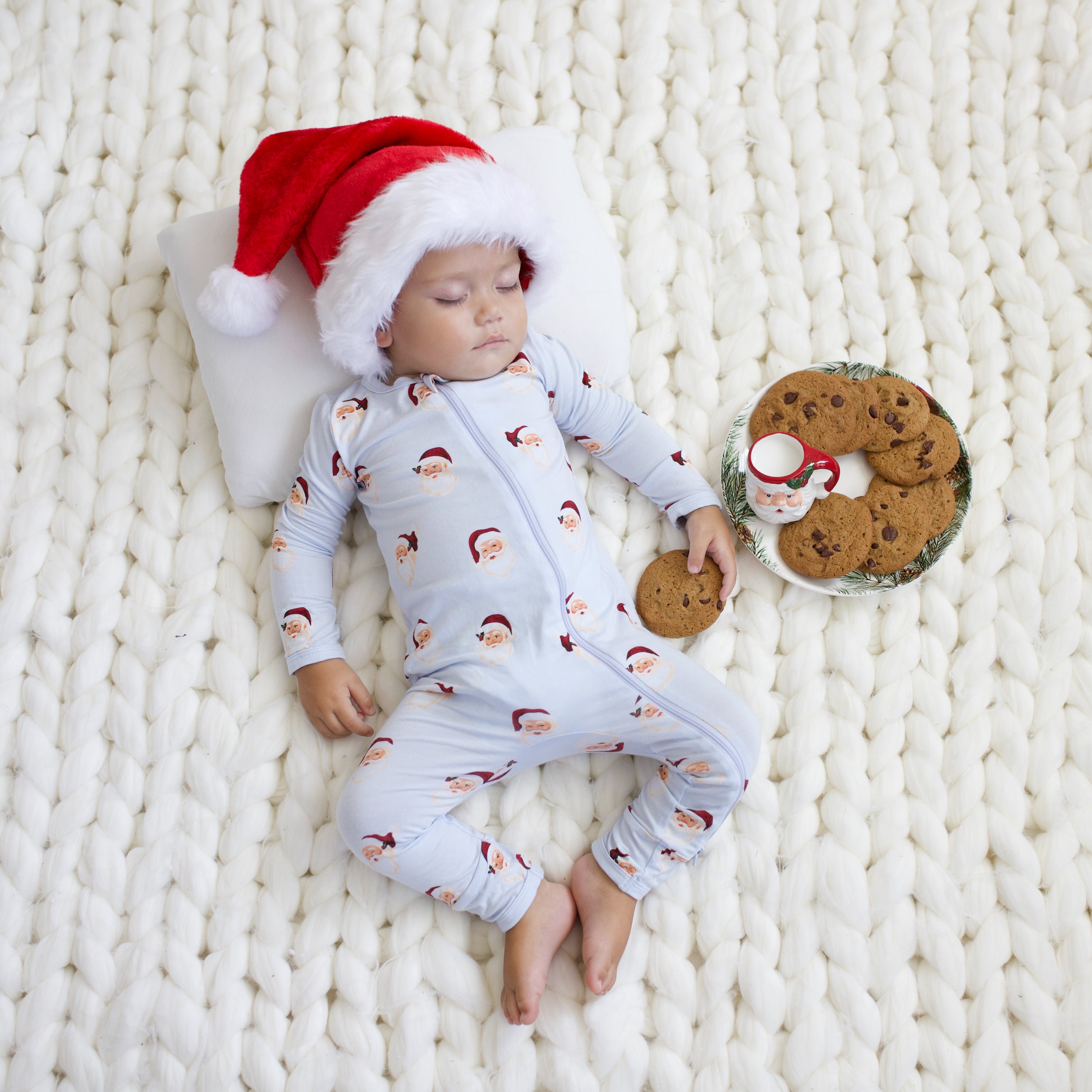 Toddler sleeping on a chunky knit cream blanket wearing the Zippered Romper in Vintage Santa and a santa hat holding a cookie with a plate of cookies beside him