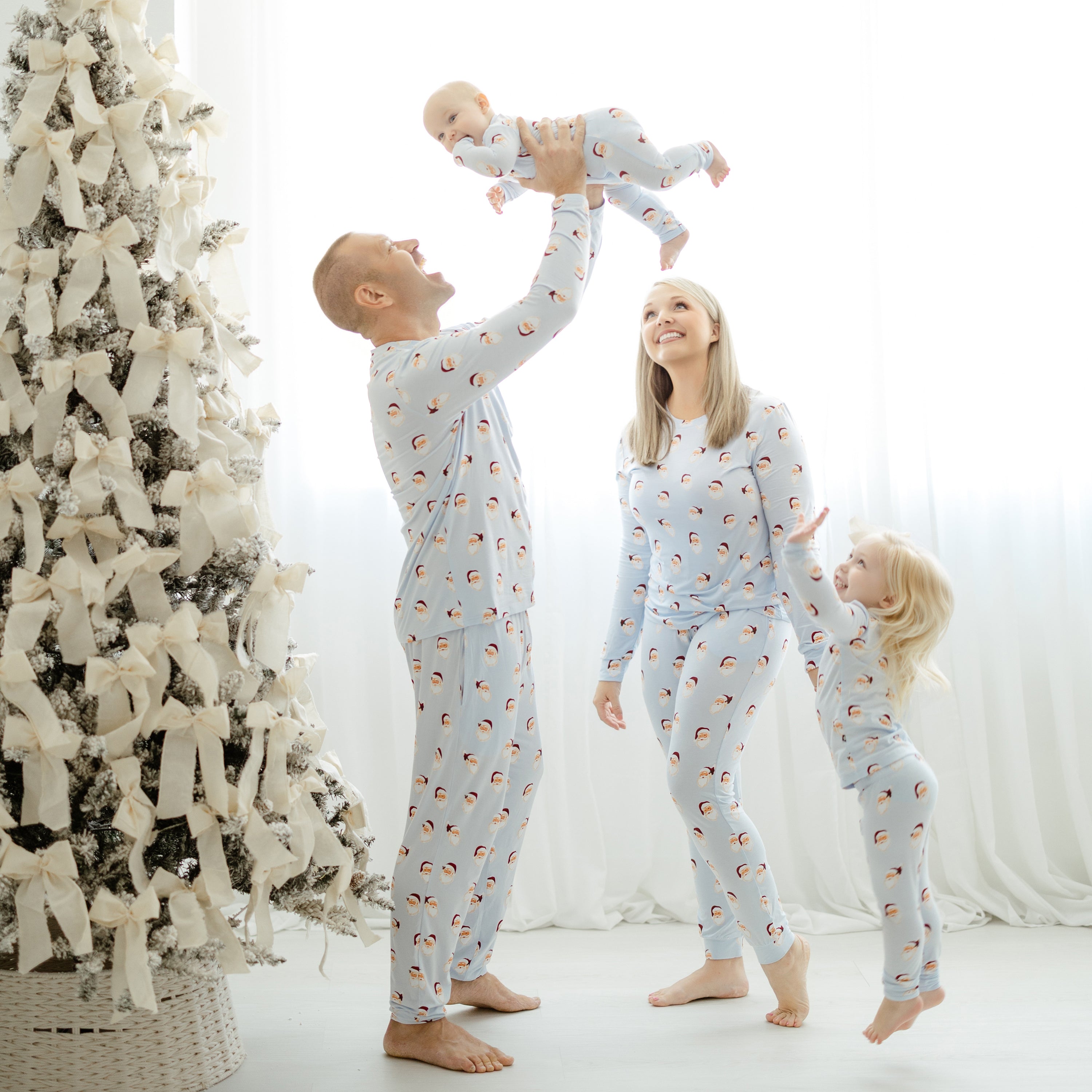 Family of four standing beside a decorated Christmas trees with white bows all matching in pajamas in the holiday print Vintage Santa