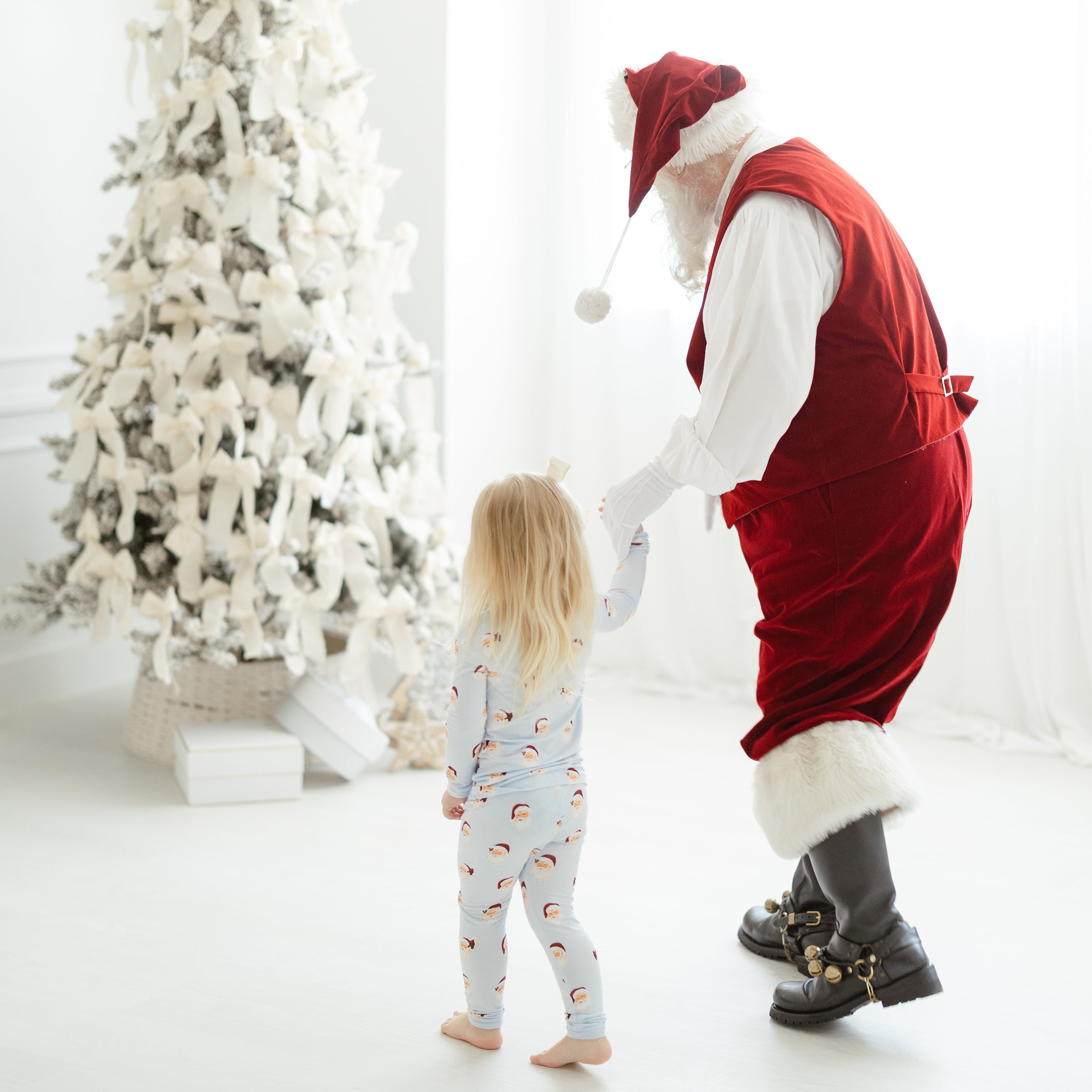 Young girl wearing the Long Sleeve Pajamas in Vintage Santa and a santa clause hold hands and walking towards a decorated Christmas tree