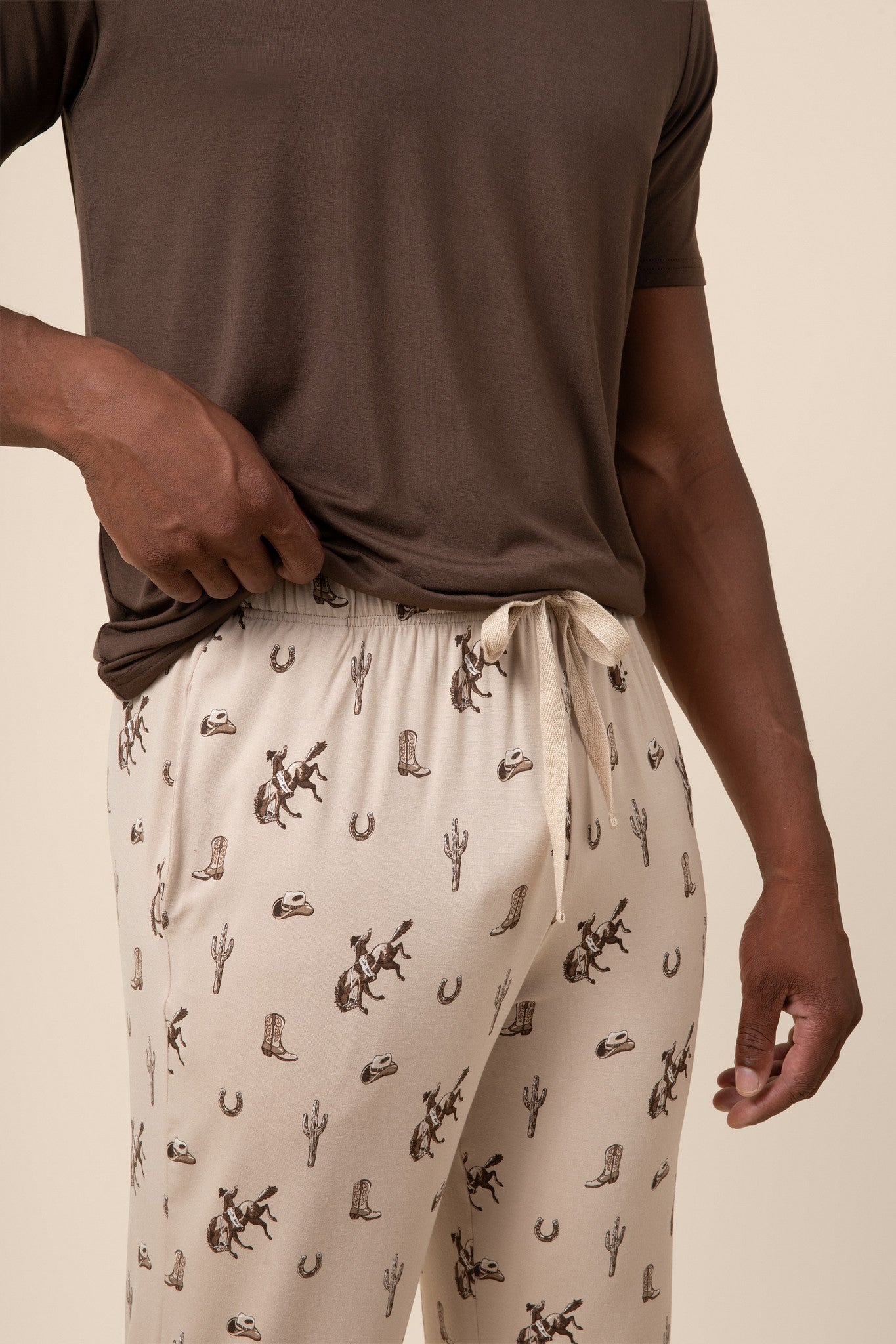 Person wearing a brown t-shirt and beige pajama pants with a cowboy, cactus , horse pattern on a beige background
