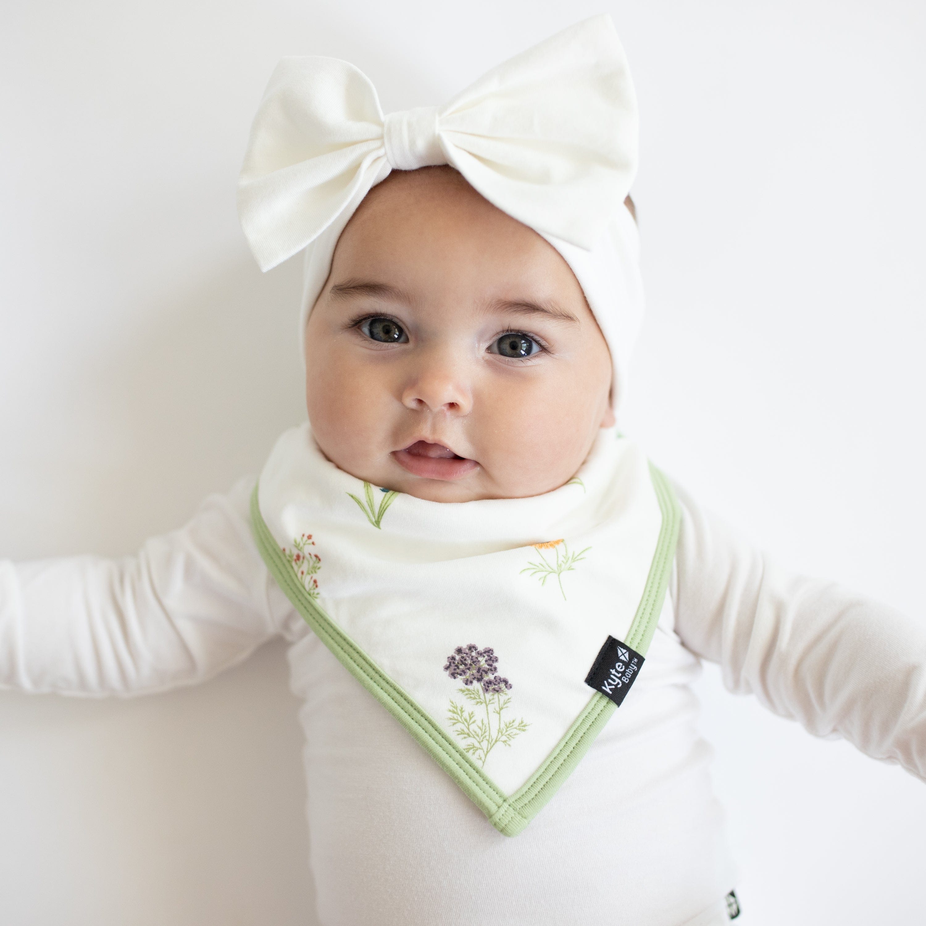 Infant wearing Bib in Wildflower over white Long Sleeve Bodysuit with Bow Headband