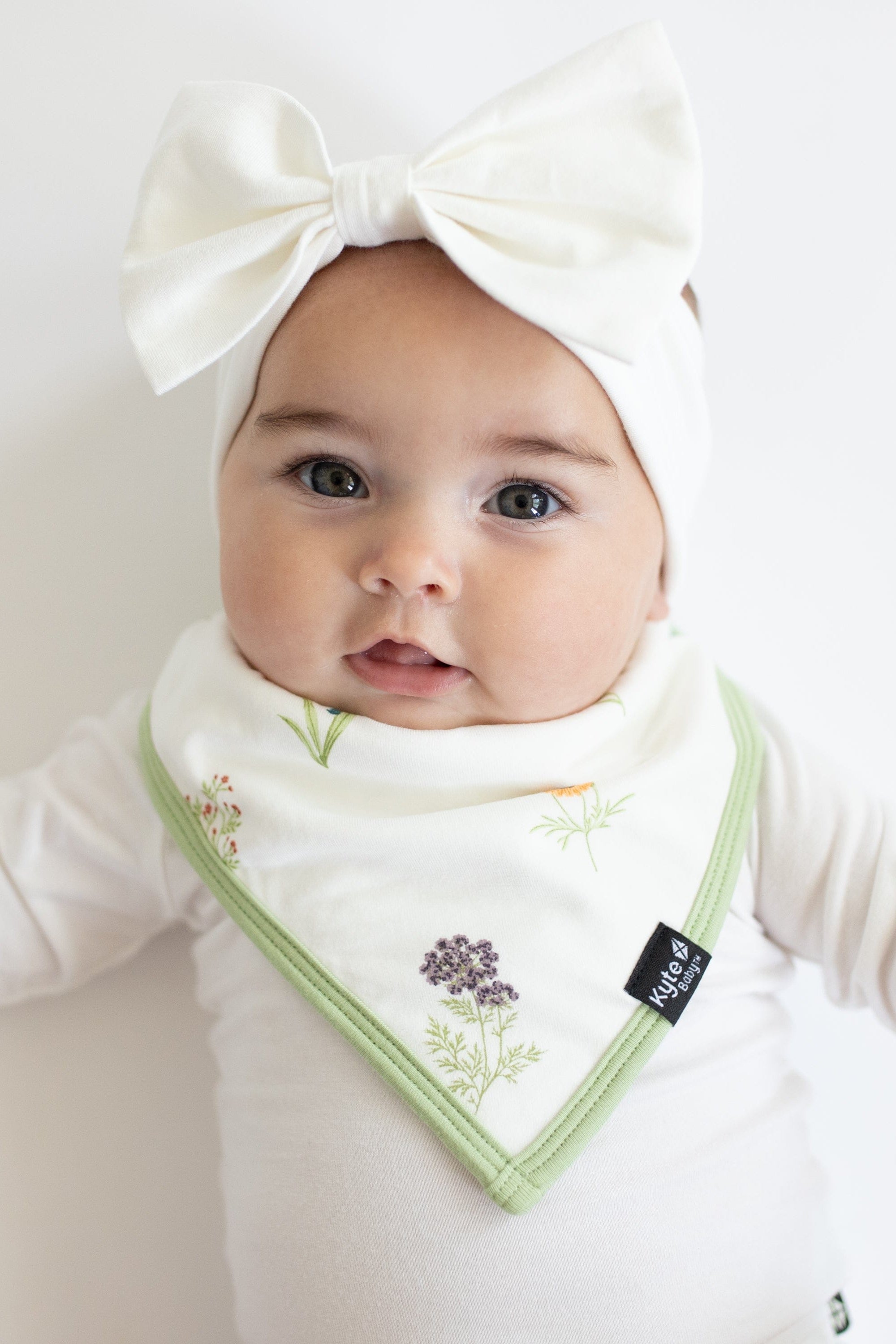Infant wearing Bib in Wildflower over white Long Sleeve Bodysuit with Bow Headband
