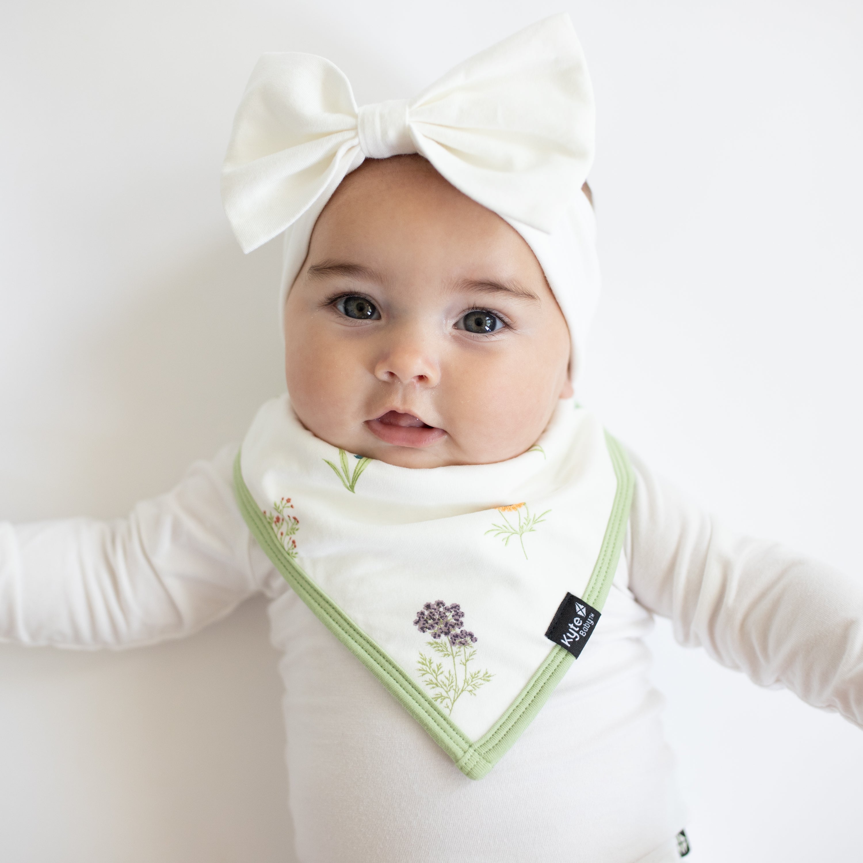 Infant wearing Bib in Wildflower over white Long Sleeve Bodysuit with Bow Headband