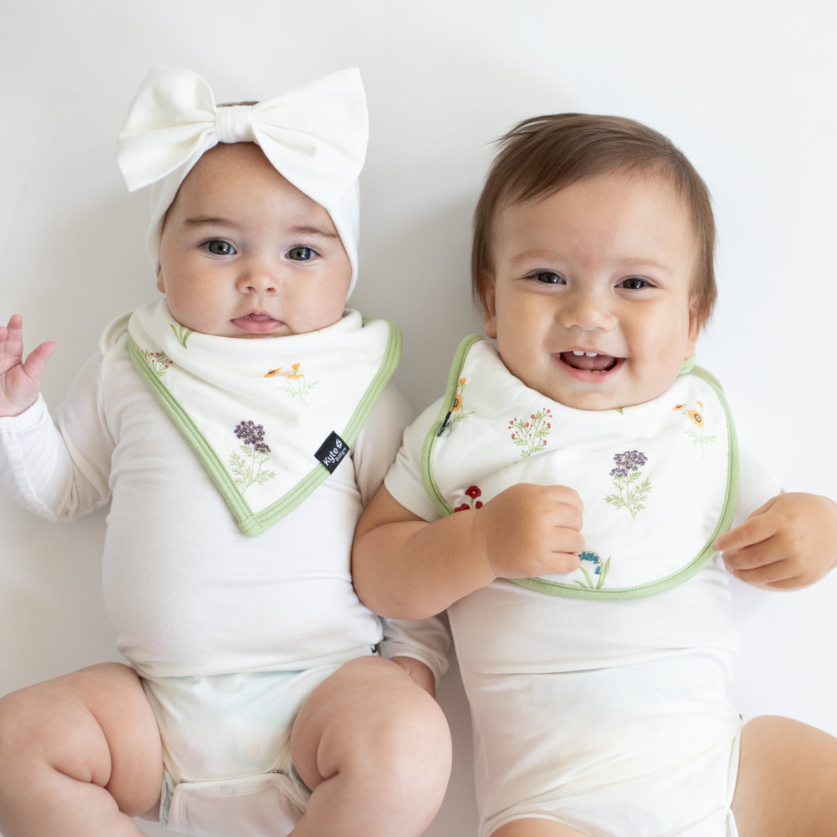 Two infant models laying side by side both wearing Cloud Bodysuits with one infant in a Bib in Wildflower and the other in a Cushy Bib in Wildflower