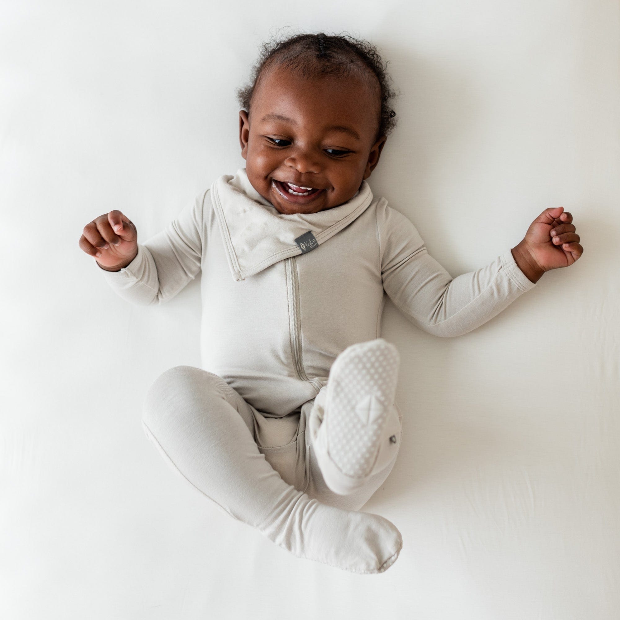Smiling infant wearing Kyte Baby Bib in Oat and matching zipper footie