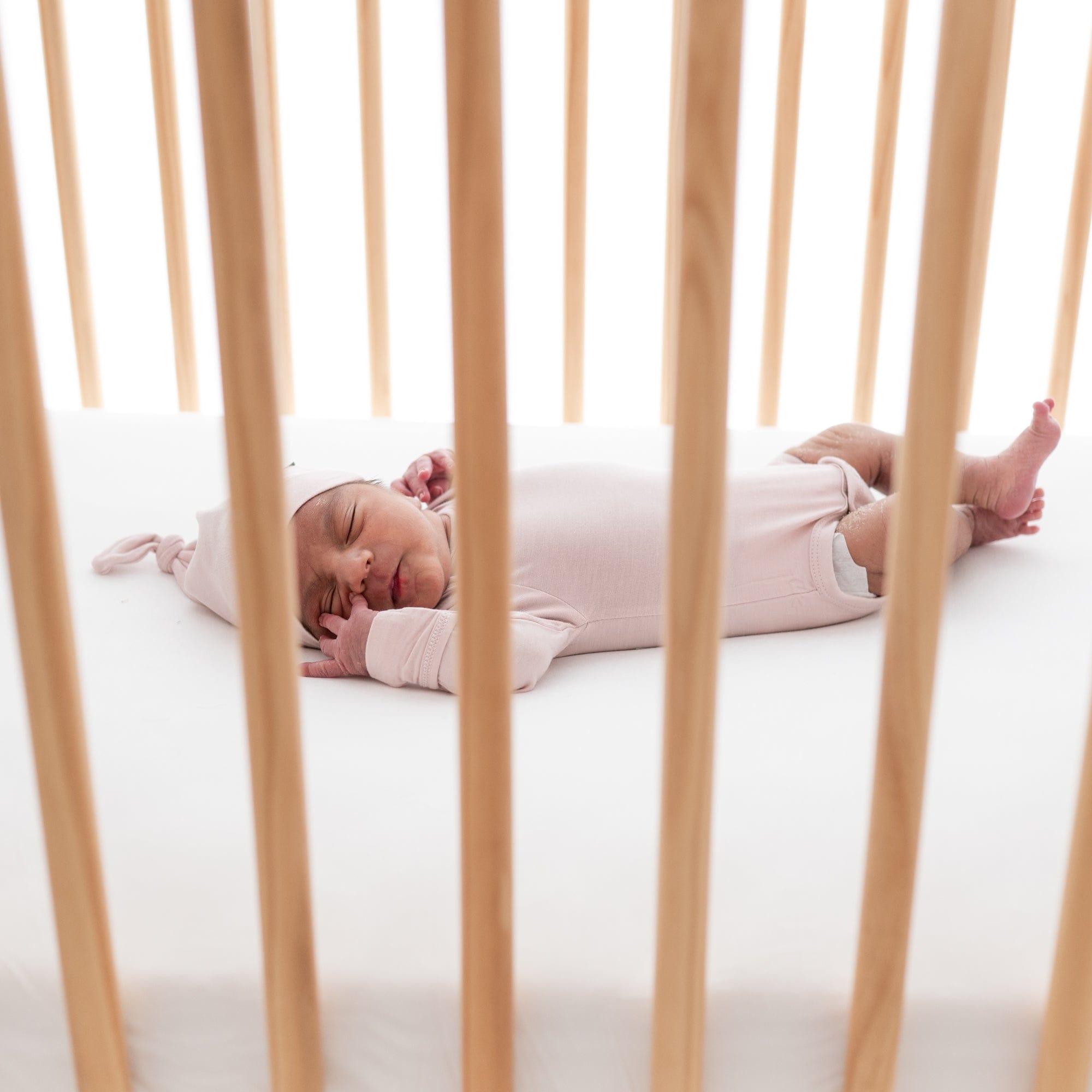 Newborn sleeping in a wooden crib while wearing a knotted Cap and long sleeve bodysuit in Blush