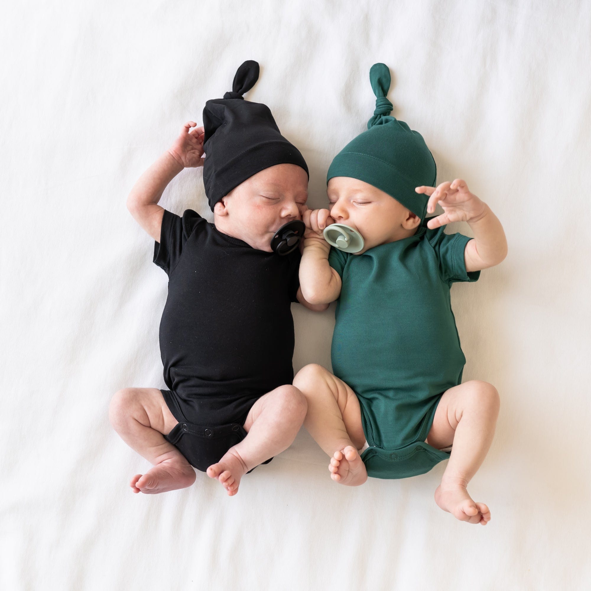 Newborns sleeping next to each other wearing Kyte Baby Knotted Caps and matching bodysuits in Midnight and Emerald