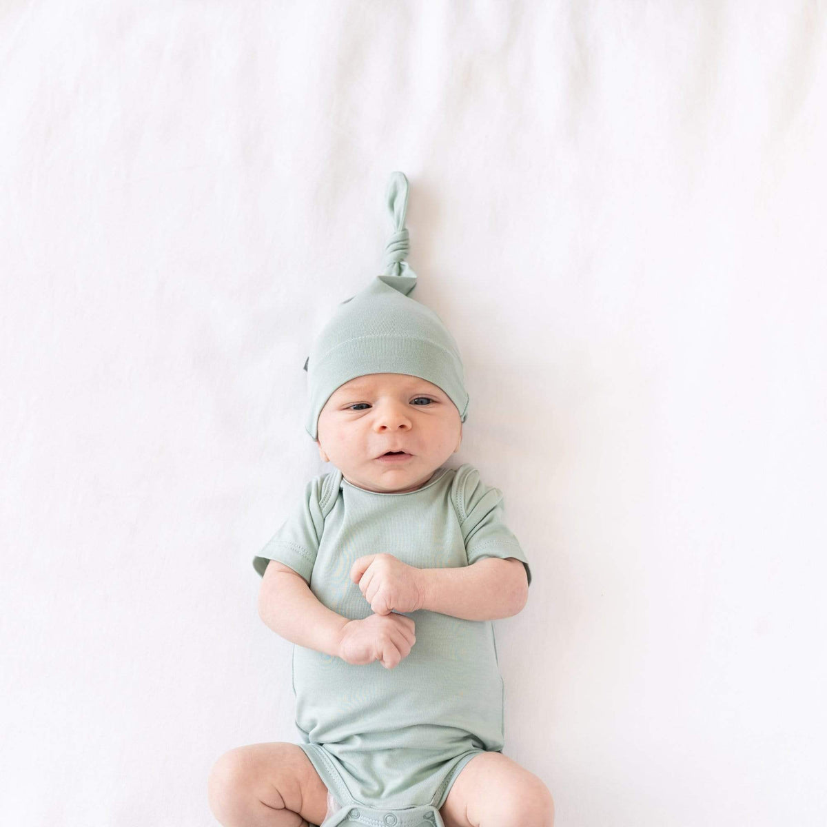 Newborn laying on a crib sheet in Oat while wearing a Bodysuit and Knotted Cap in Sage