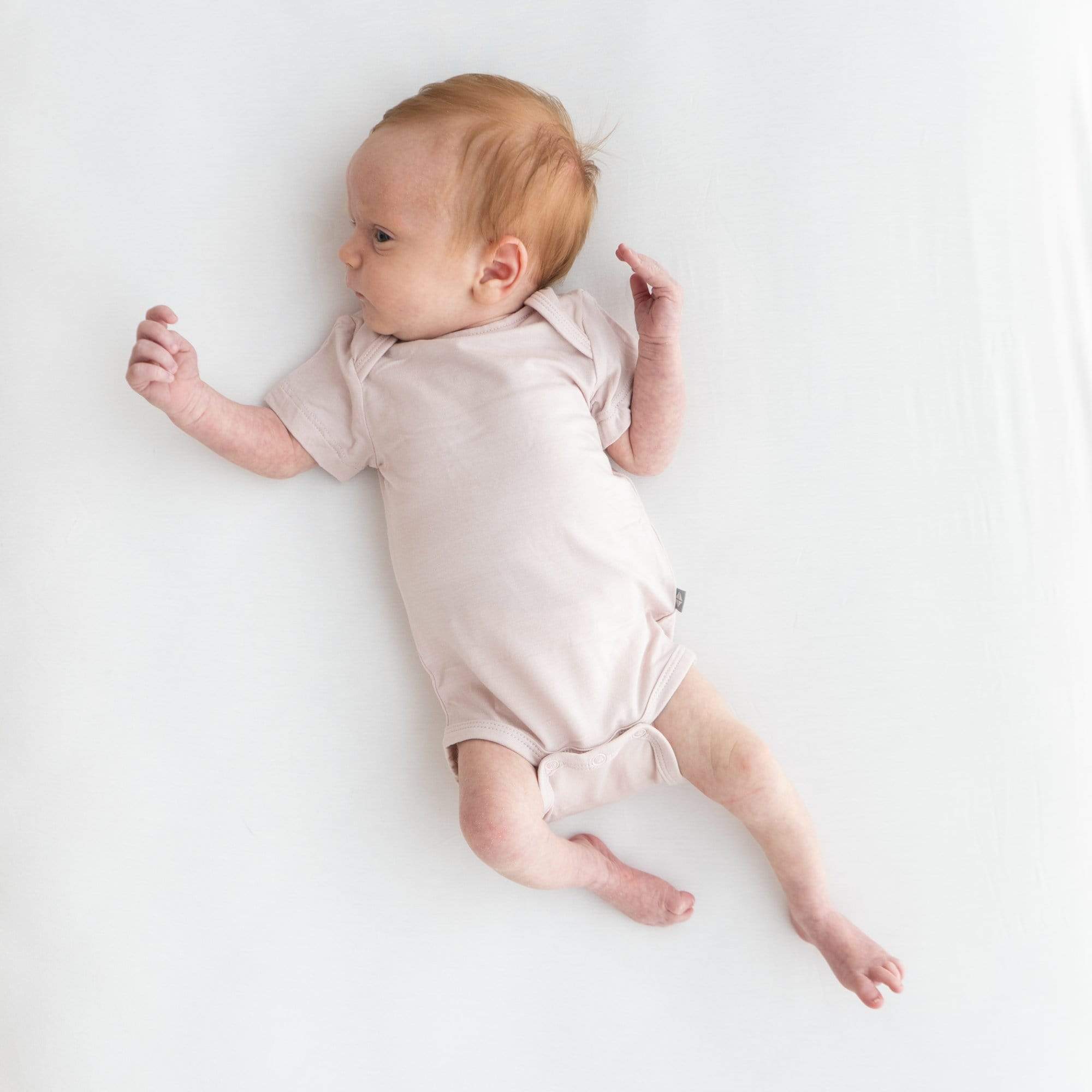 Newborn laying on a white surface, looking towards the right and wearing a Kyte Baby Bodysuit in Blush