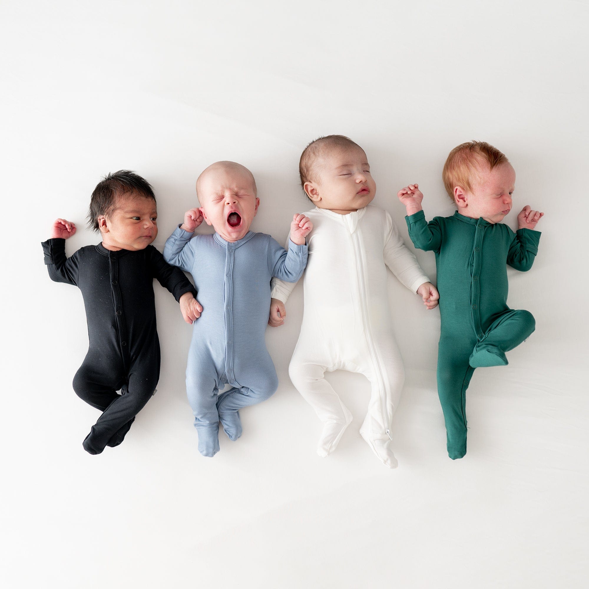 Babies laying next to each other wearing Kyte Baby footies in Midnight, Slate, Cloud and Emerald