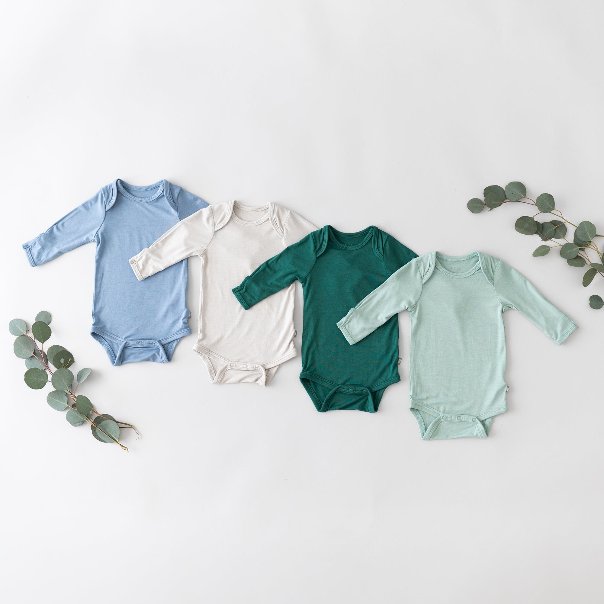 Flat lay of layered Kyte Baby Long Sleeve Bodysuits in Slate, Cloud, Emerald and Sage