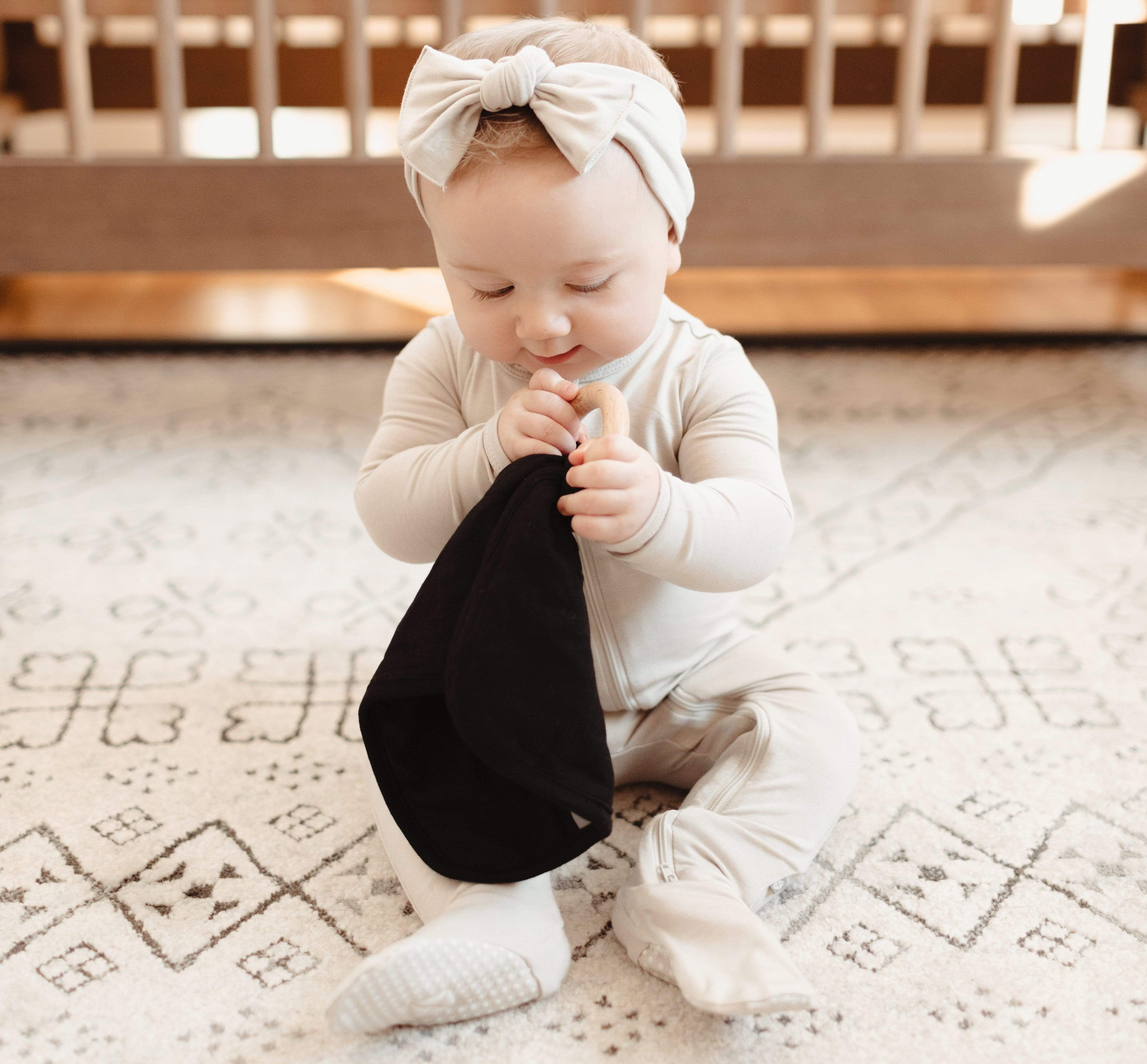 Infant playing with Kyte Baby Lovey in Midnight while wearing Zippered Footie and Headband in Oat