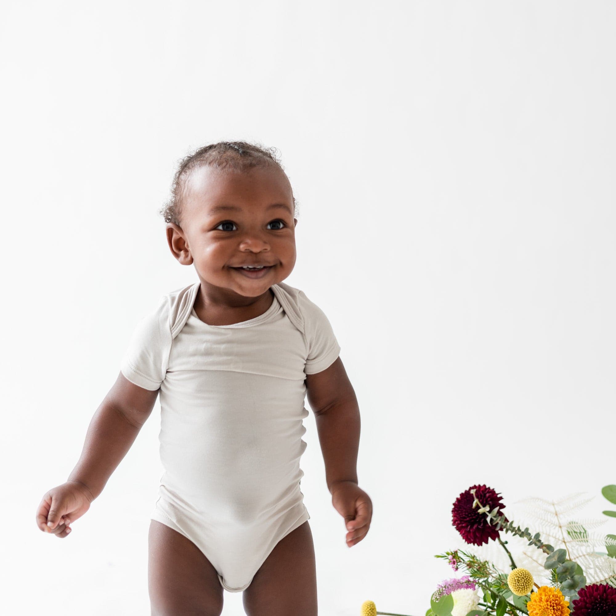 Smiling baby standing next to flowers in a Kyte Baby Bodysuit in Oat