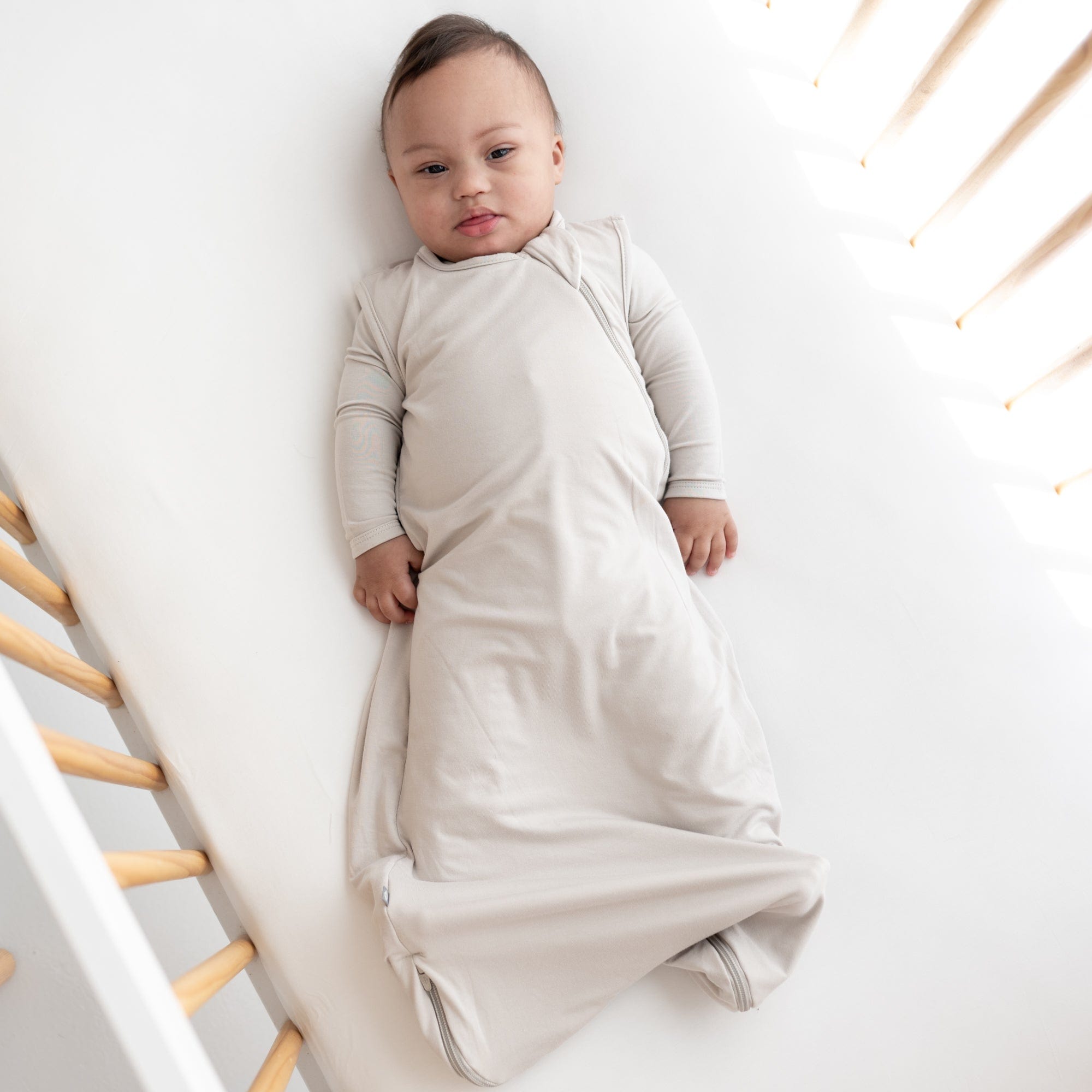 Infant laying in crib while wearing 0.5 Tog Sleep Bag in Oat with a matching long sleeve bodysuit underneath